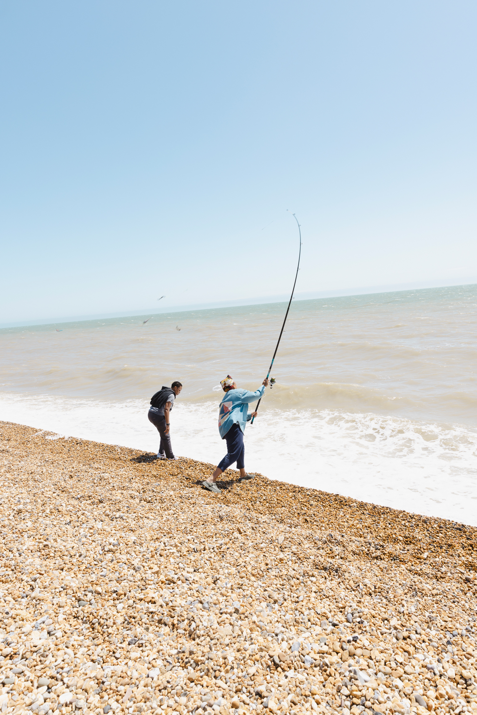 Two people fishing from pebble beach with long rod extended. White foam waves meet brown stones under pale blue sky.