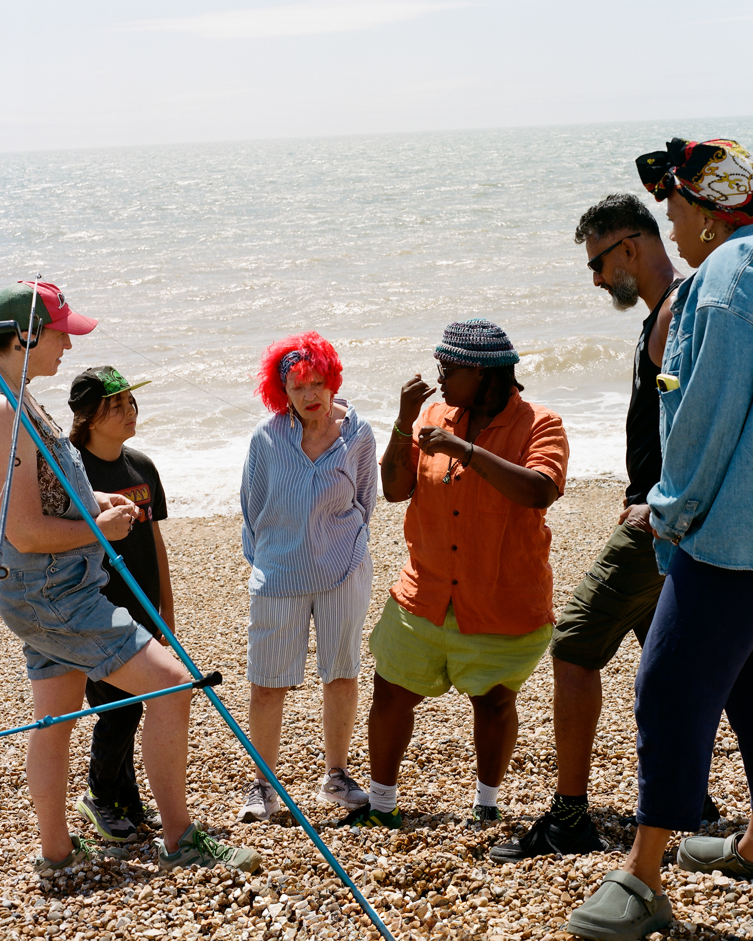 Group of people on pebble beach with sea in background. Adults and children wearing colourful clothing, one holding blue pole.