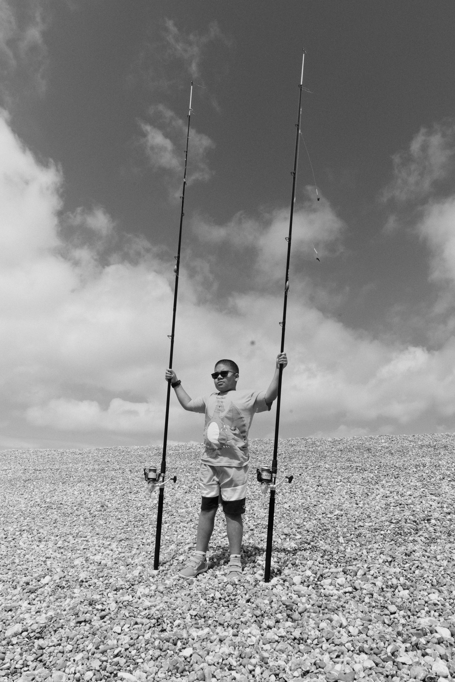 Man in sunglasses holding two fishing rods aloft on pebble beach, black and white image with cloudy sky background.