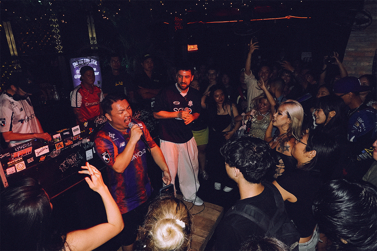 DJ performing in crowded nightclub with red and blue lighting, people dancing around DJ booth, dark interior with scattered lights.