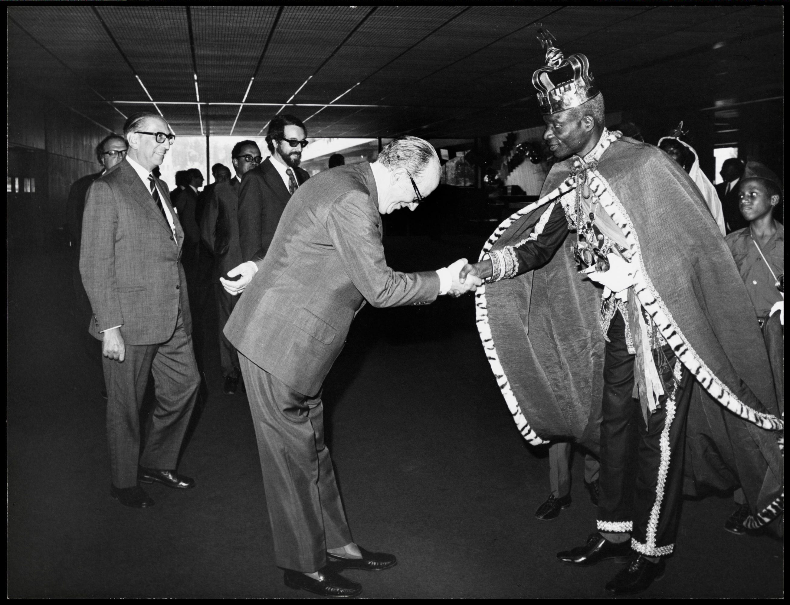 Black and white image of man in suit bowing to shake hands with person in ornate traditional robes and crown, surrounded by officials.