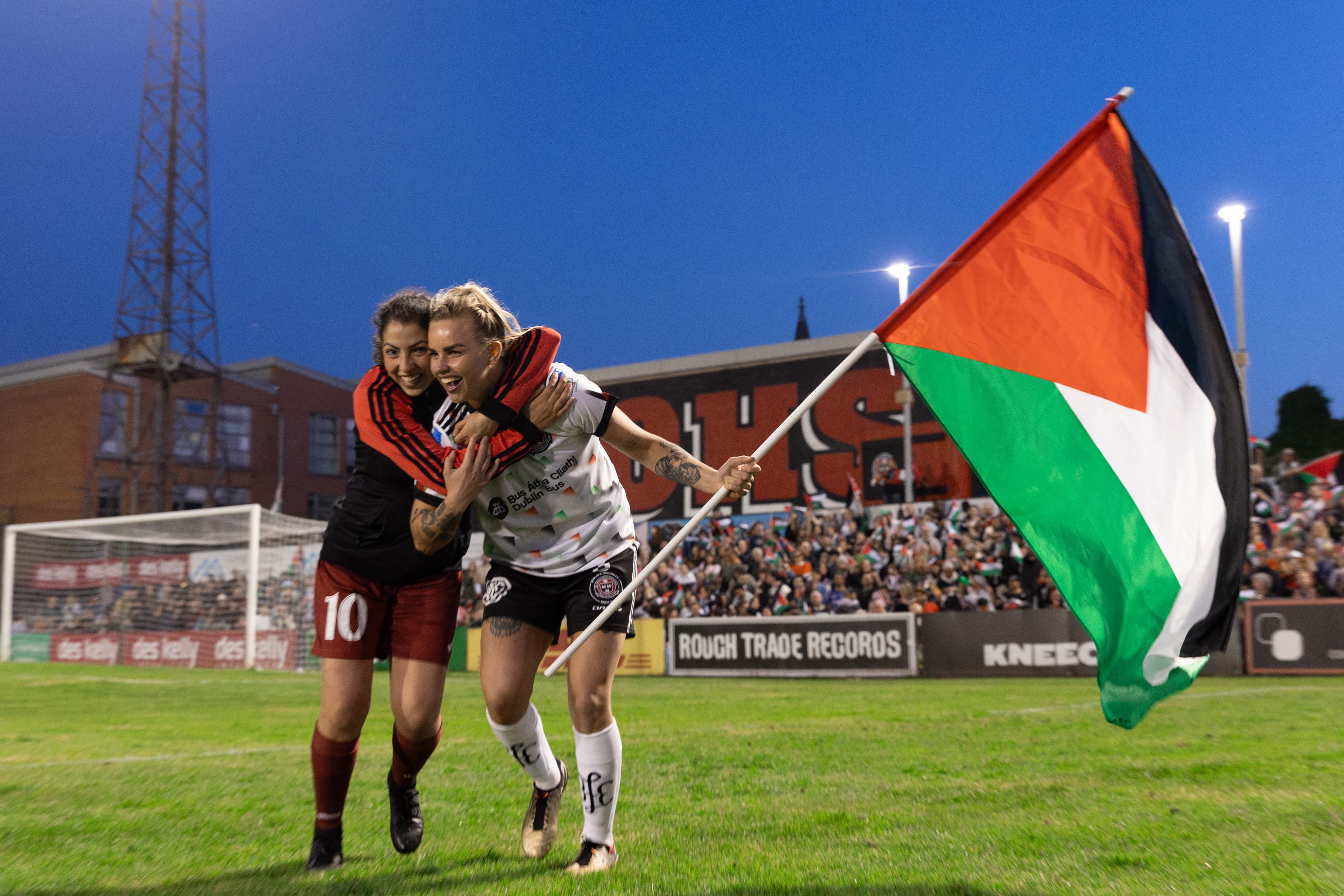 Two men embracing while holding a Palestinian flag on a football pitch.