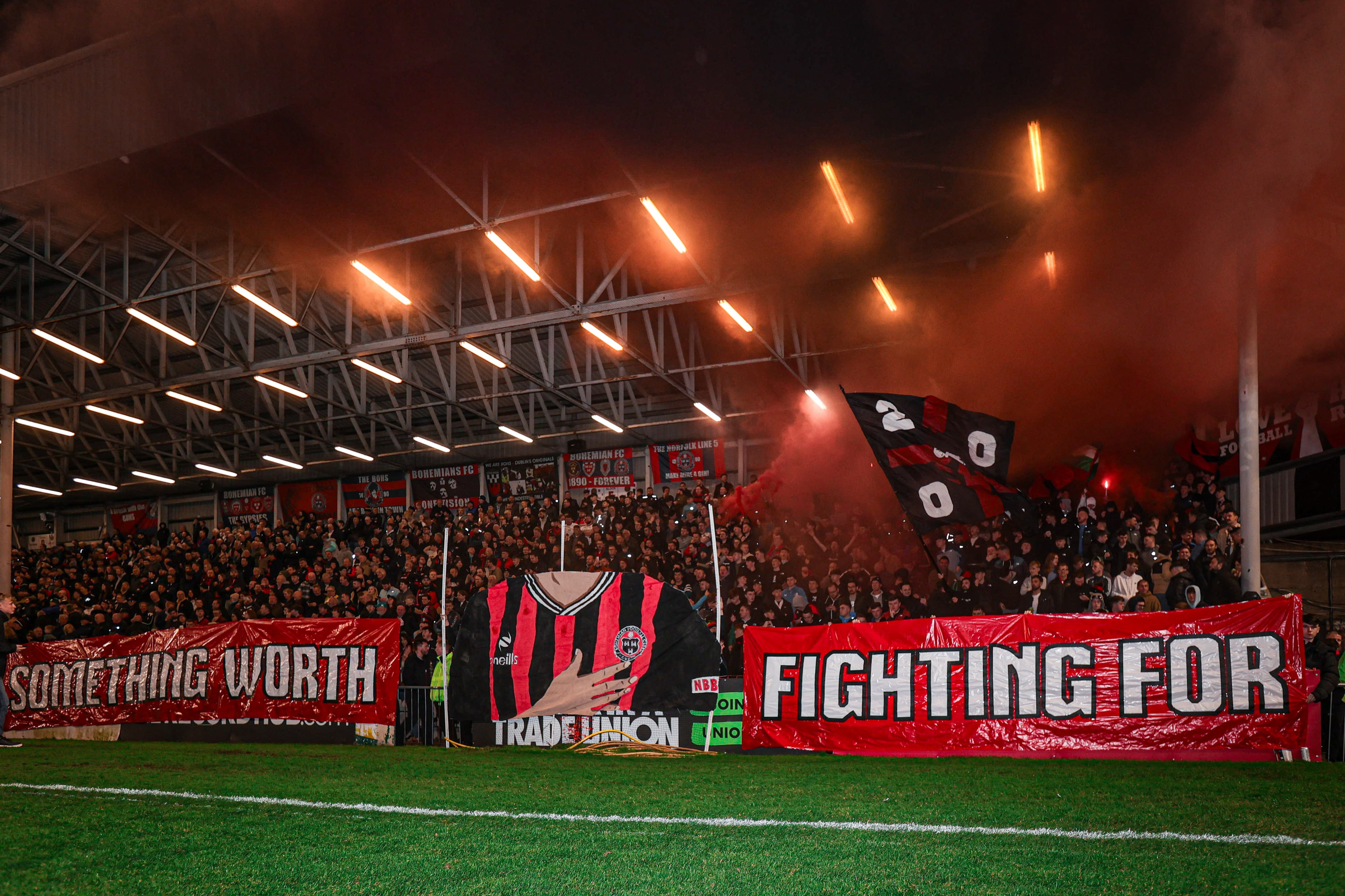 Crowded football stadium at night, red-and-black banners with slogans, floodlights illuminating the scene.