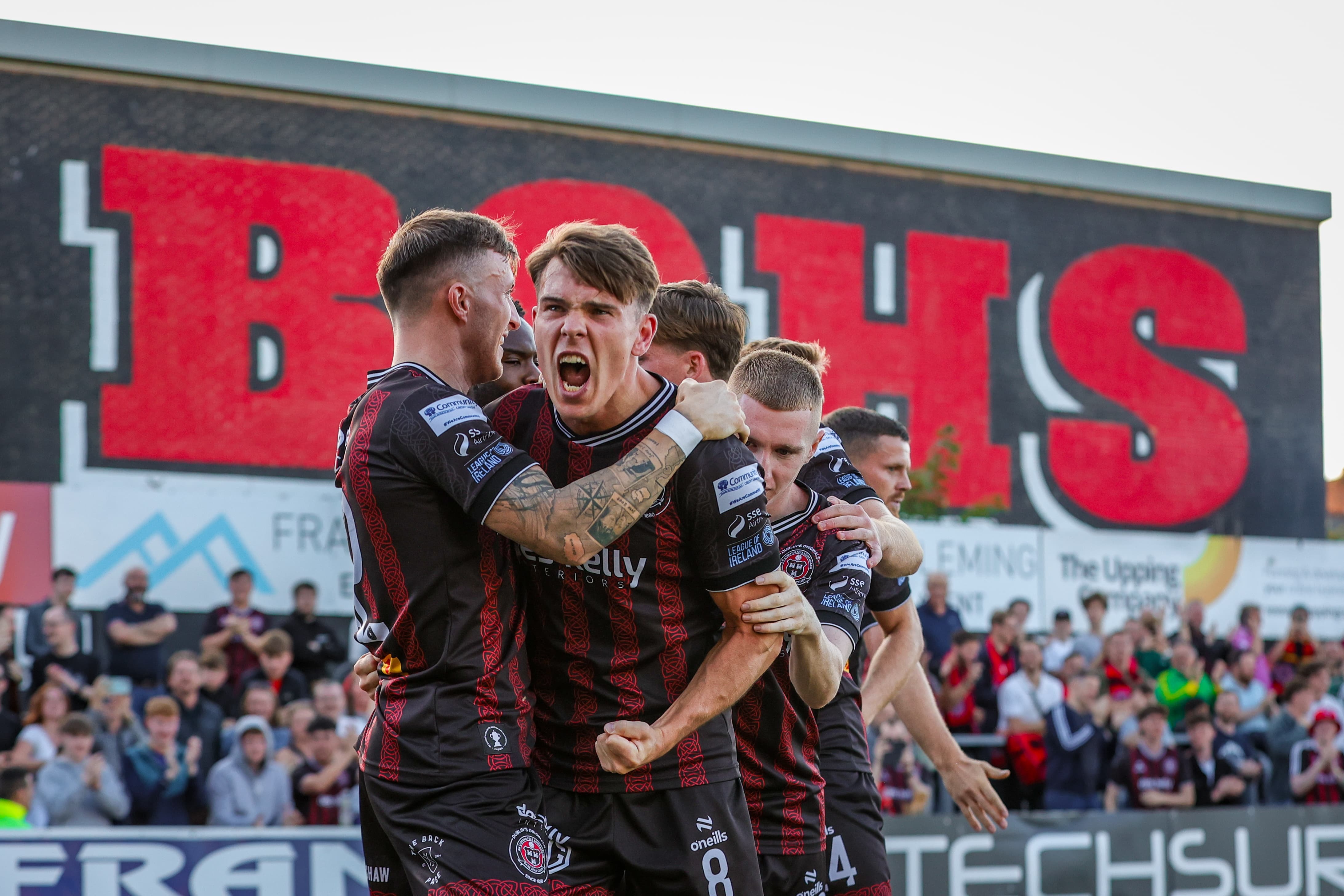 A group of men celebrating on a football pitch, with a large "IBBO'S" sign in the background.