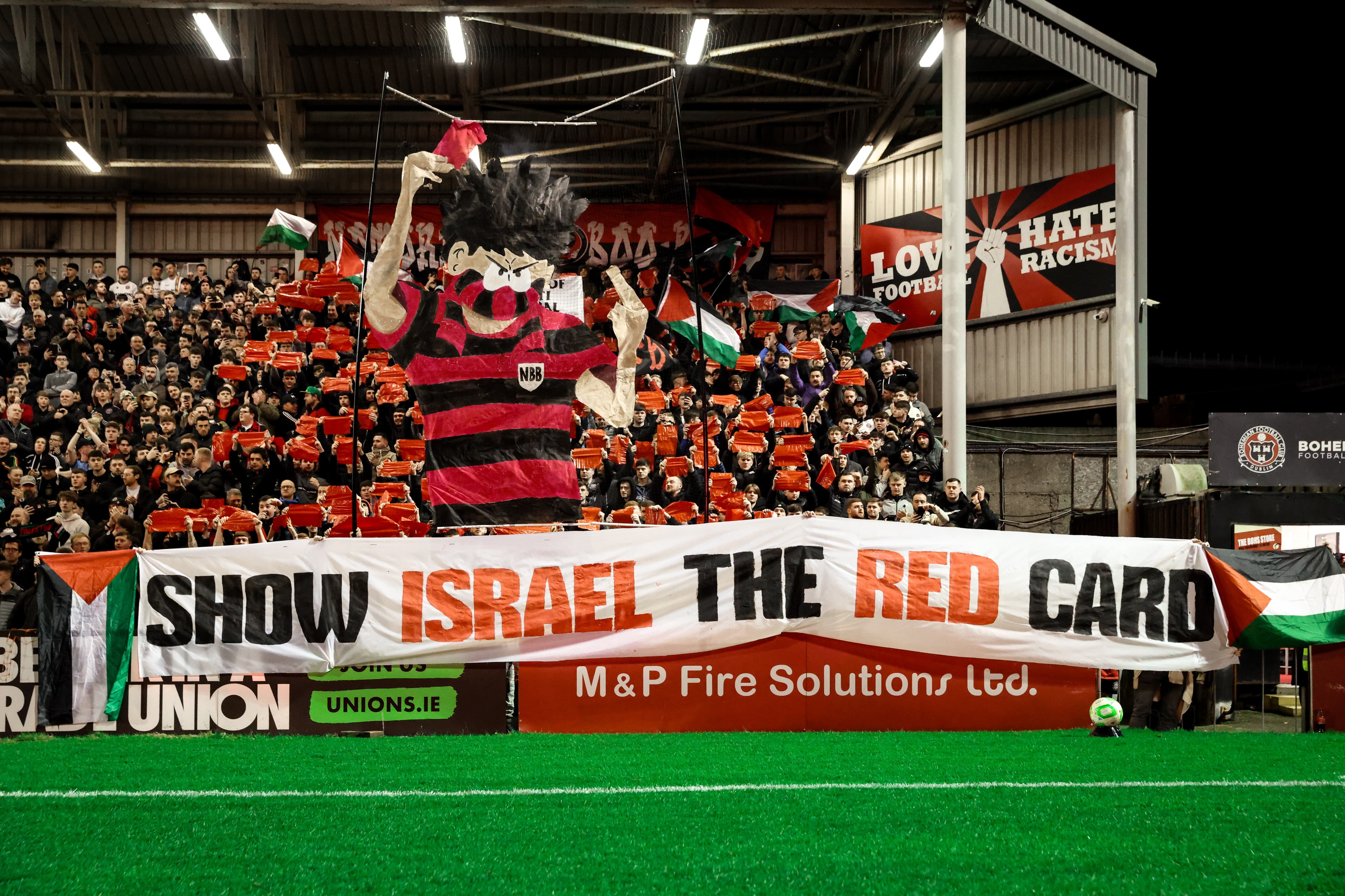 Enthusiastic football fans in a stadium, holding red and white flags and banners, including one that says "Show Israel the red card".