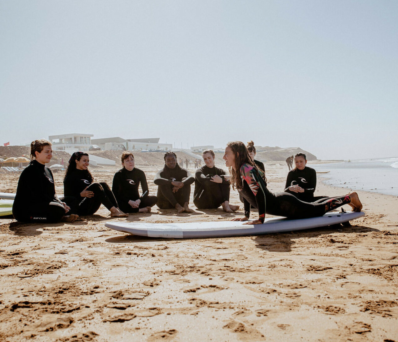 Group of surfers sitting on surfboard on beach with buildings in background.