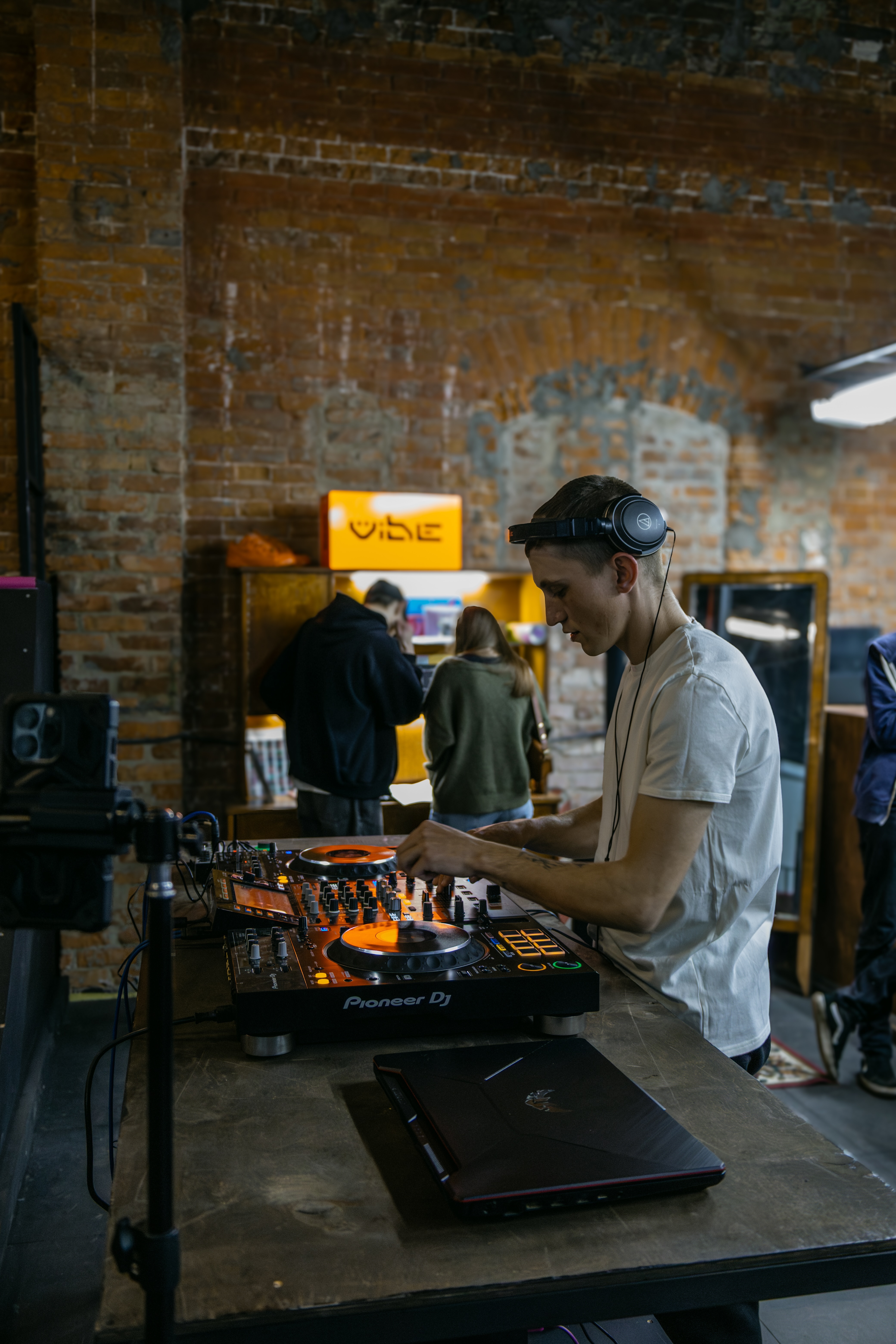 Man wearing headphones DJ-ing at a soundboard in a music venue with brick walls.