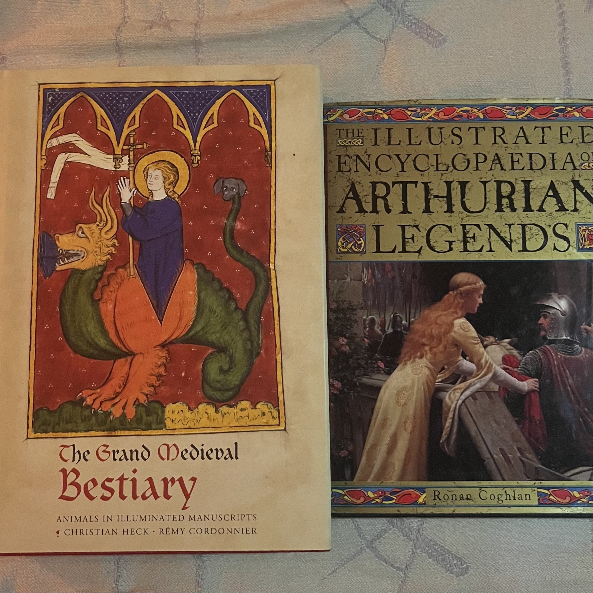 Two books: "The Grand Medieval Bestiary" with illuminated manuscript cover showing figure in blue robes within red arched frame, and "Illustrated Encyclopaedia of Arthurian Legends" with medieval scene cover.