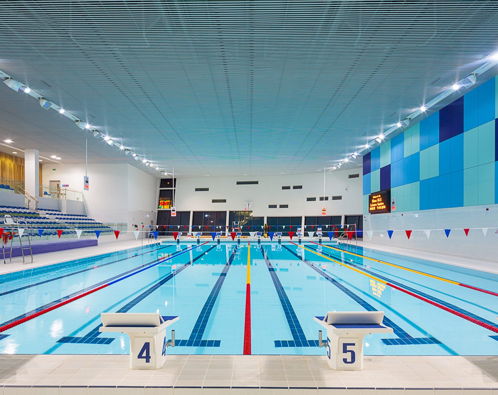 Indoor swimming pool with multiple lanes, blue water, starting blocks numbered 4 and 5, tiered seating, and geometric blue wall panels.