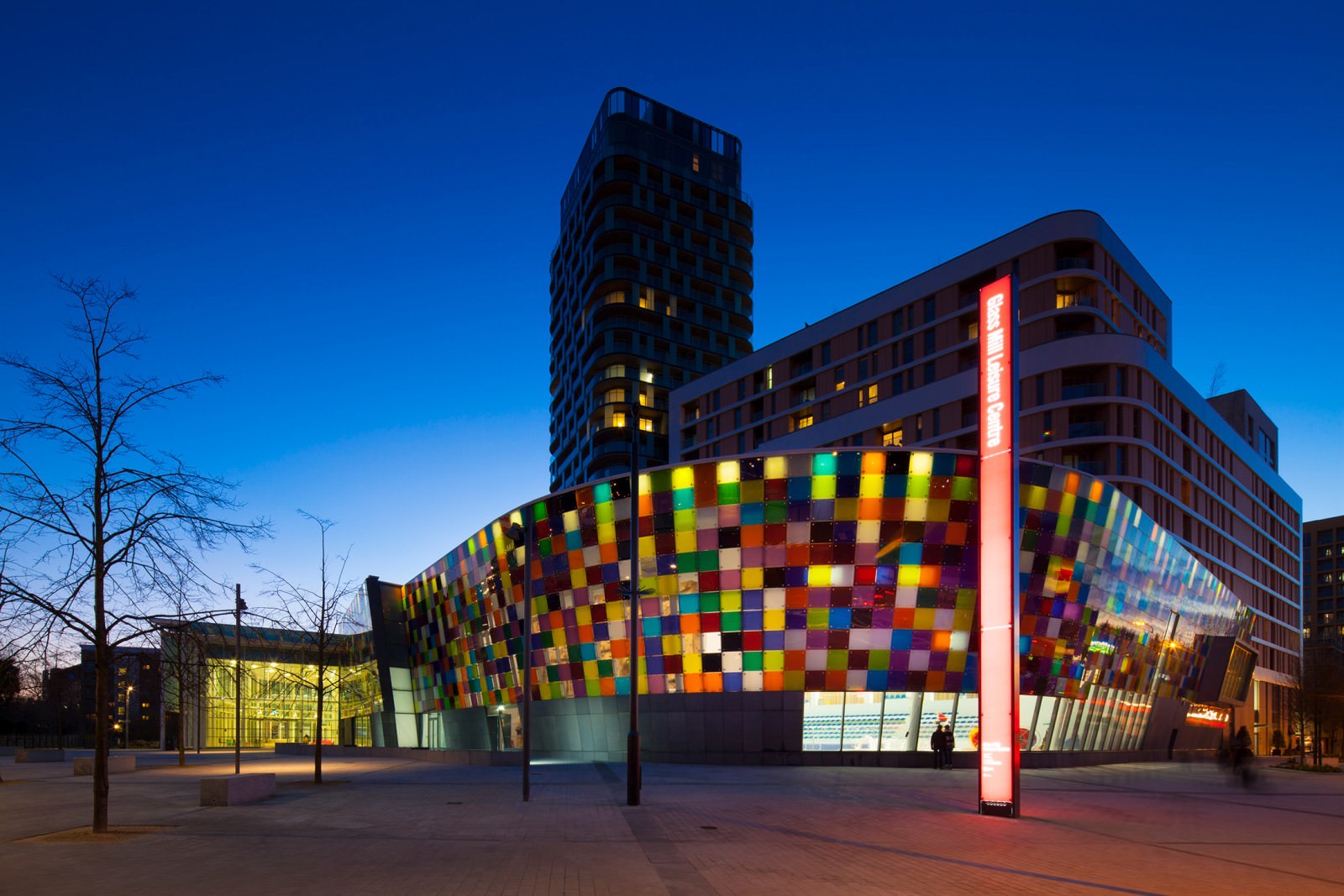 Modern building with multicoloured mosaic facade in geometric pattern, curved glass sections, and tall tower block behind against blue evening sky.