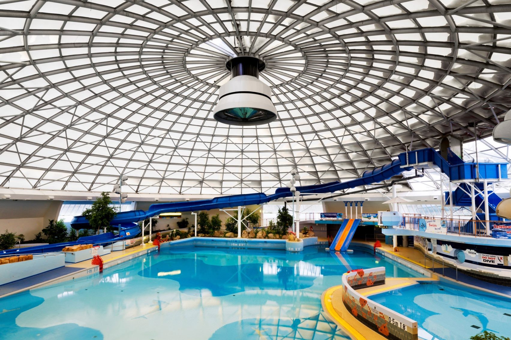 Indoor swimming pool with blue water slides under large white domed glass roof with geometric grid pattern and central circular opening.