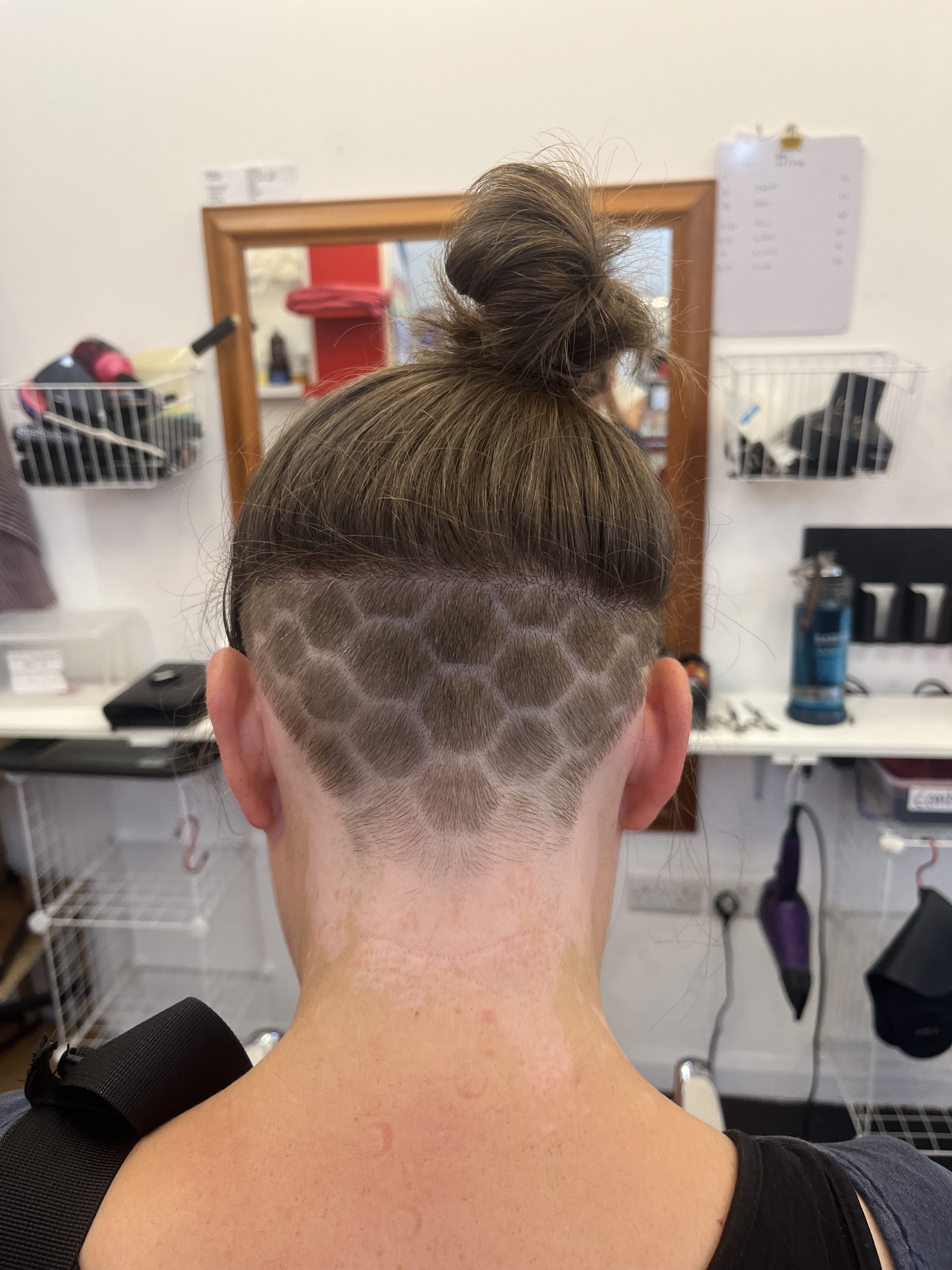 Back of person's head with shaved pattern and topknot hairstyle in salon setting.