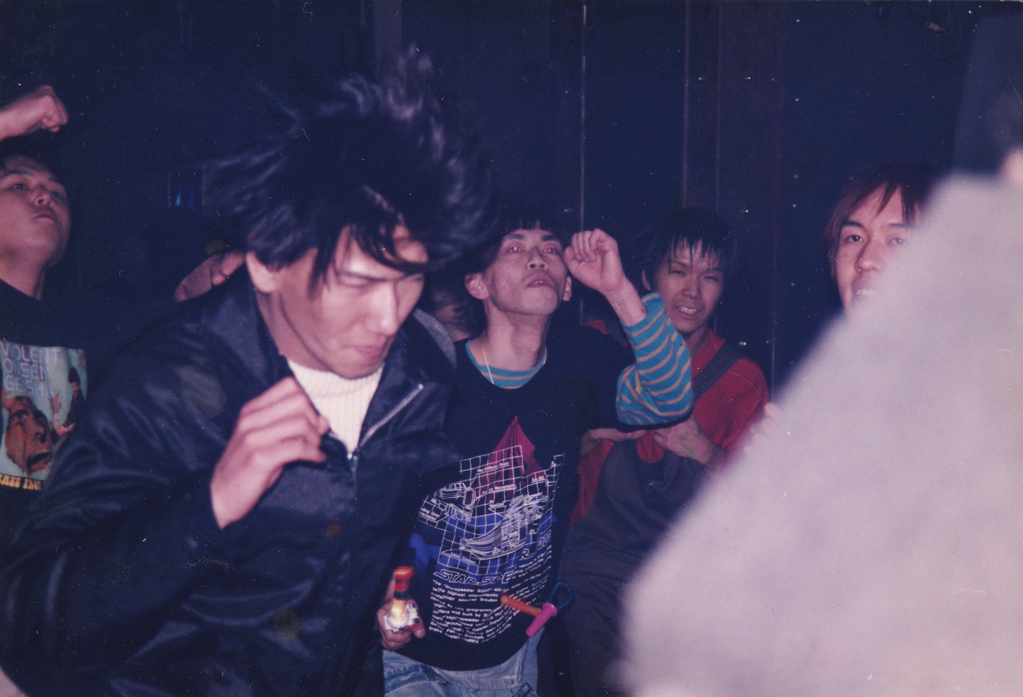 Group of people at indoor gathering, dark lighting with pink/purple tones, some wearing striped clothing and leather jackets.