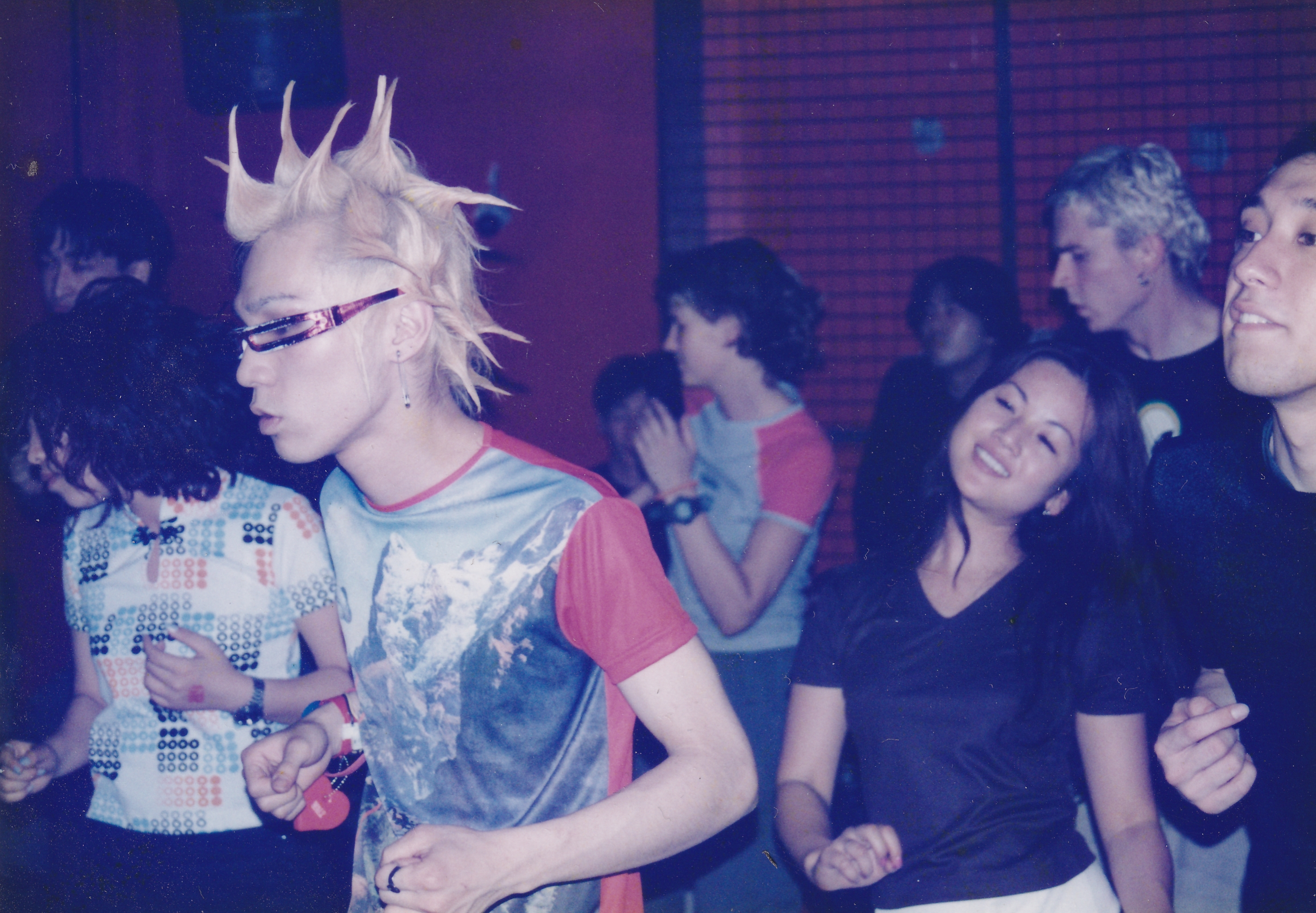 Person with spiked white hair and sunglasses in colourful patterned shirt amongst crowd in purple-lit indoor venue.