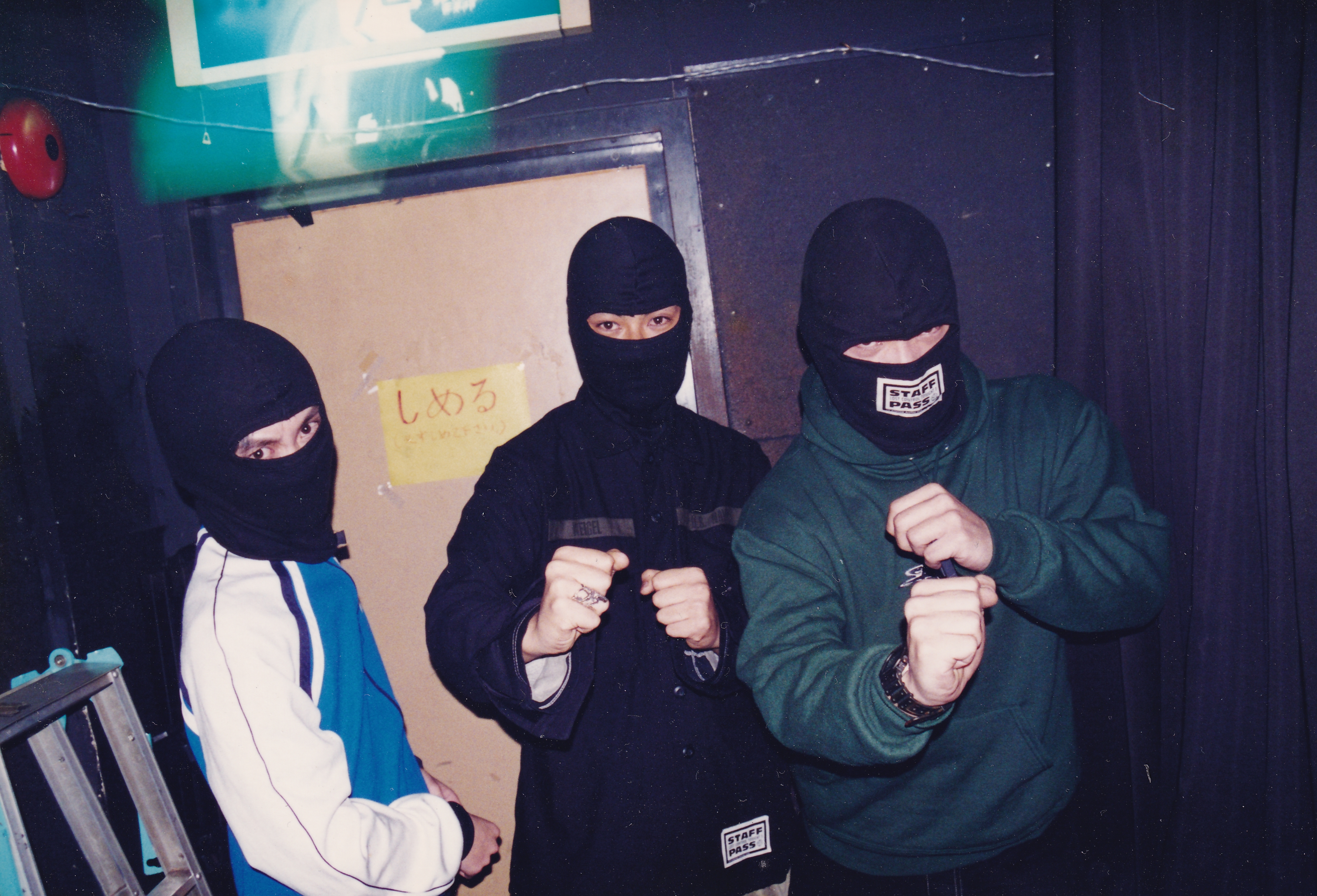 Three people wearing black balaclavas indoors, one in white tracksuit with blue stripes, one in black top, one in green jumper.