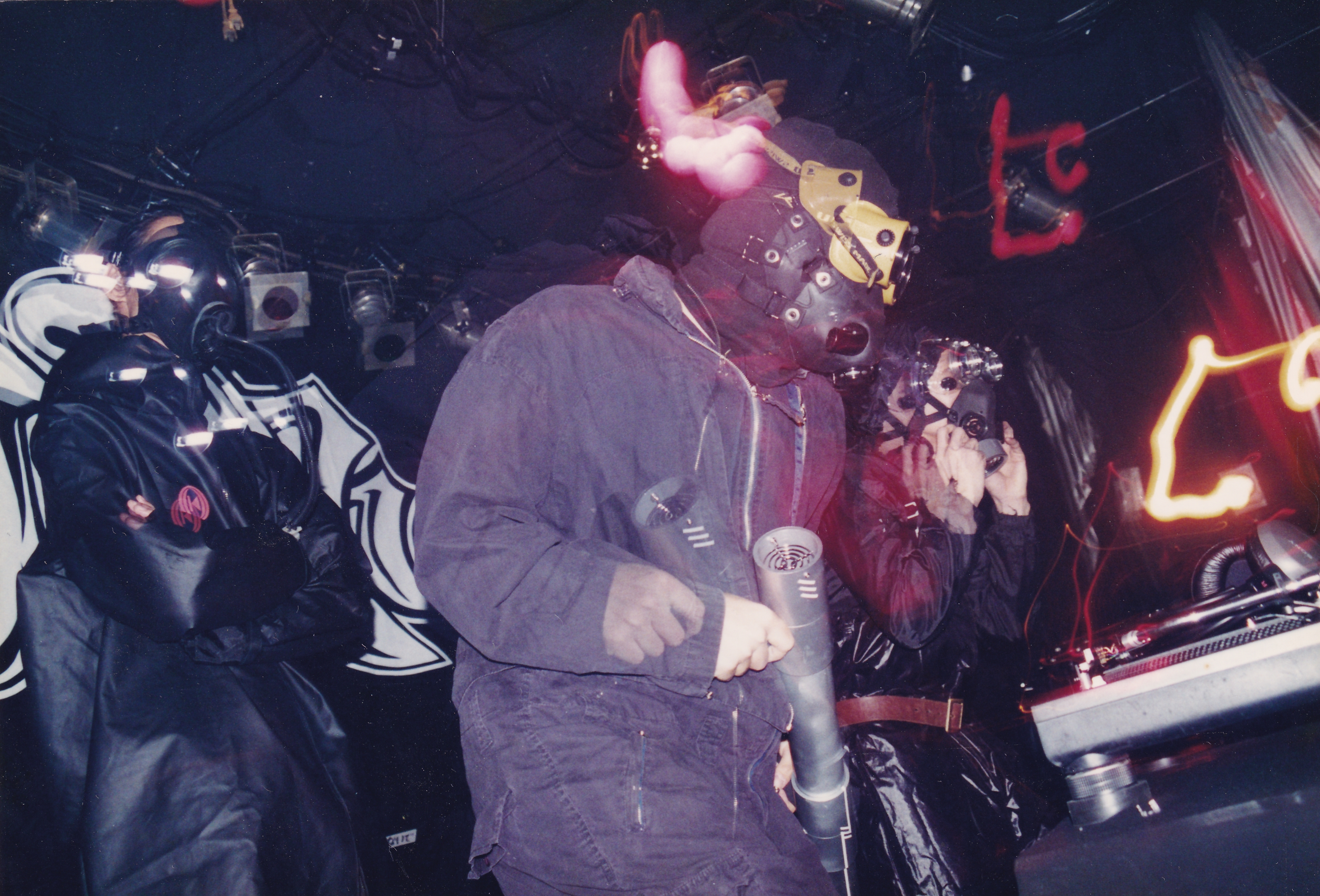 DJ wearing animal mask performing at nightclub with red lighting, crowd visible in background, electronic music equipment present.