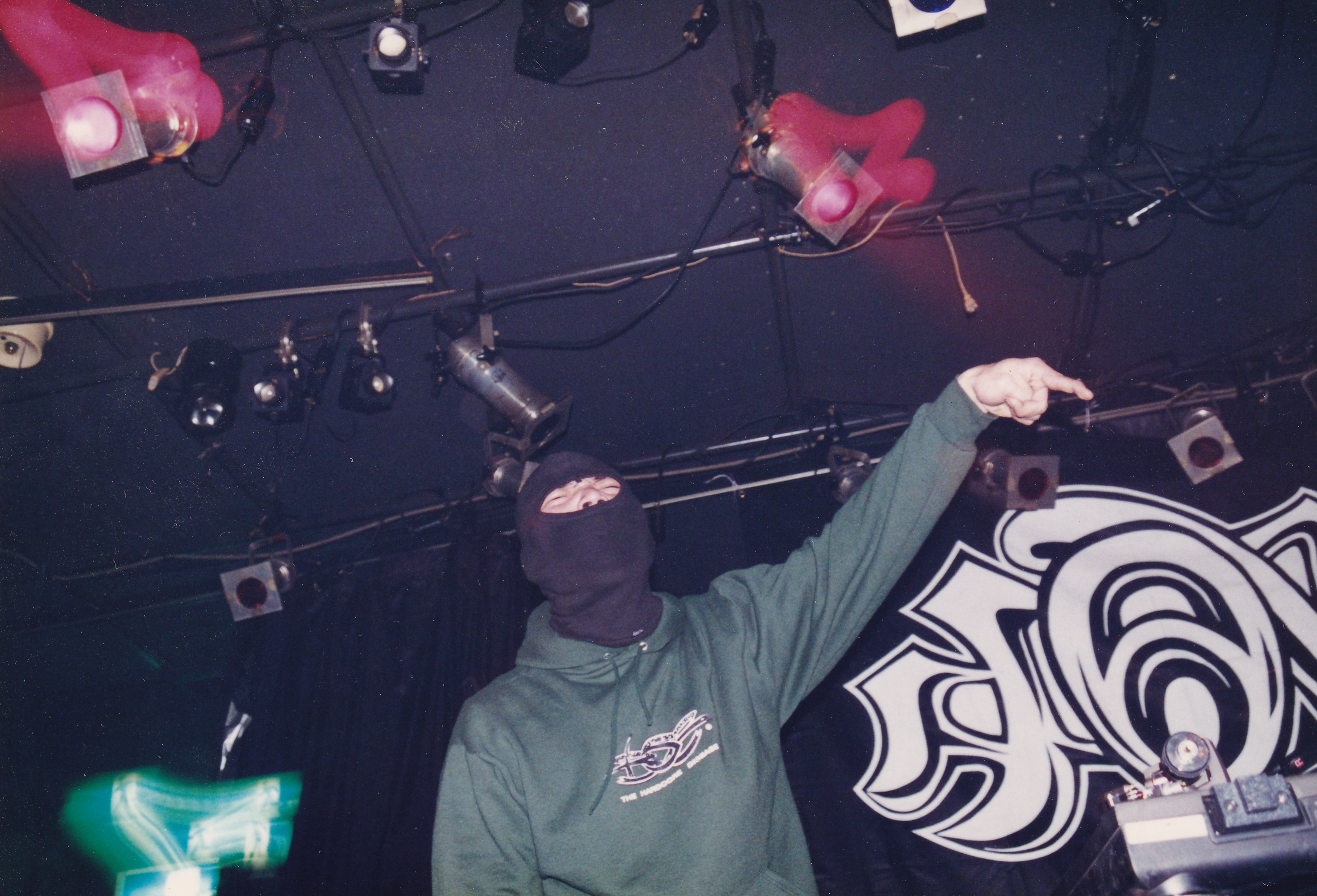 Overhead view of DJ performing at decks with green hoodie, purple lighting, and pink/red stage lights visible.