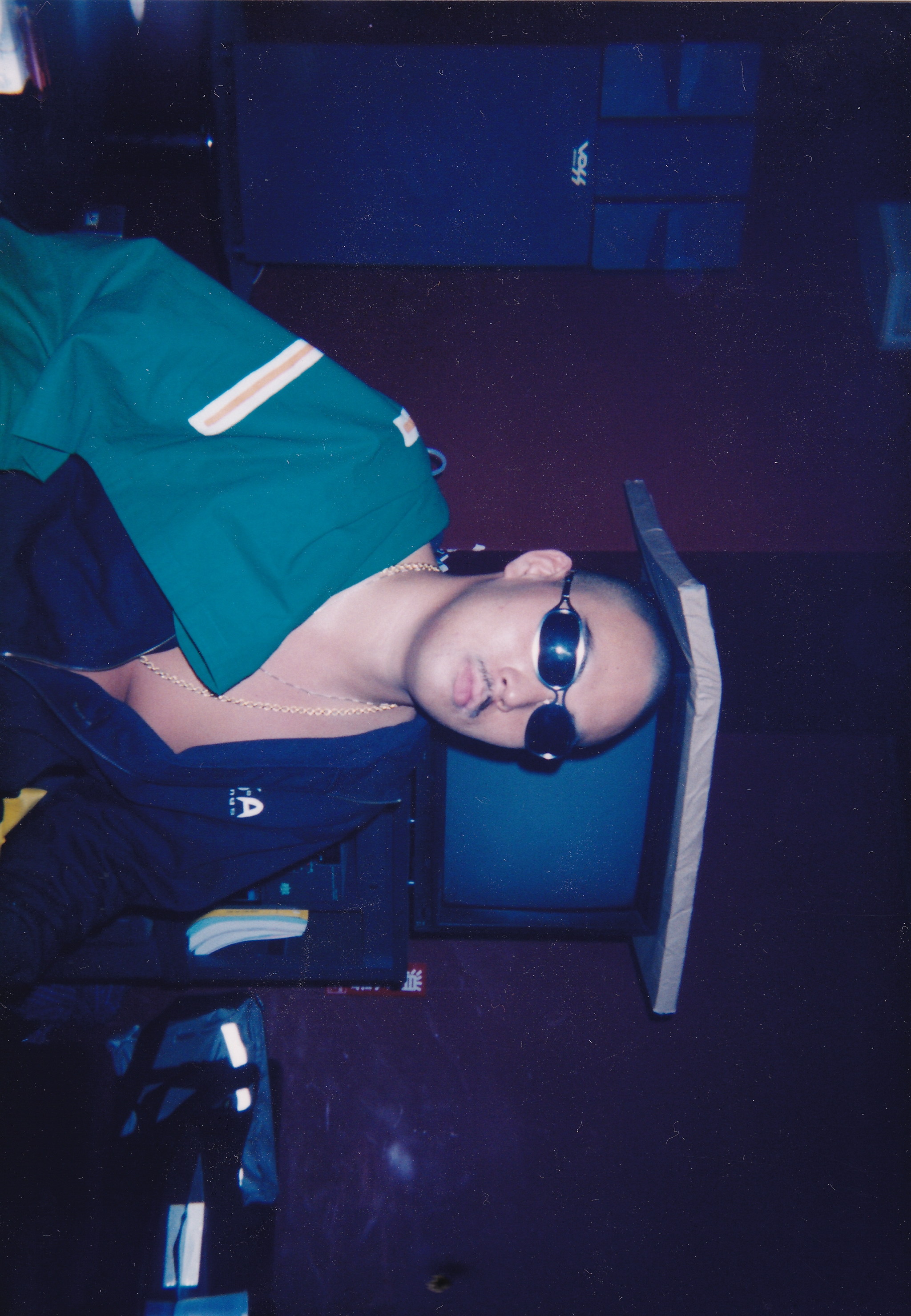 Person with dark sunglasses and teal jumper lying on aircraft seat in dim blue-lit cabin interior.