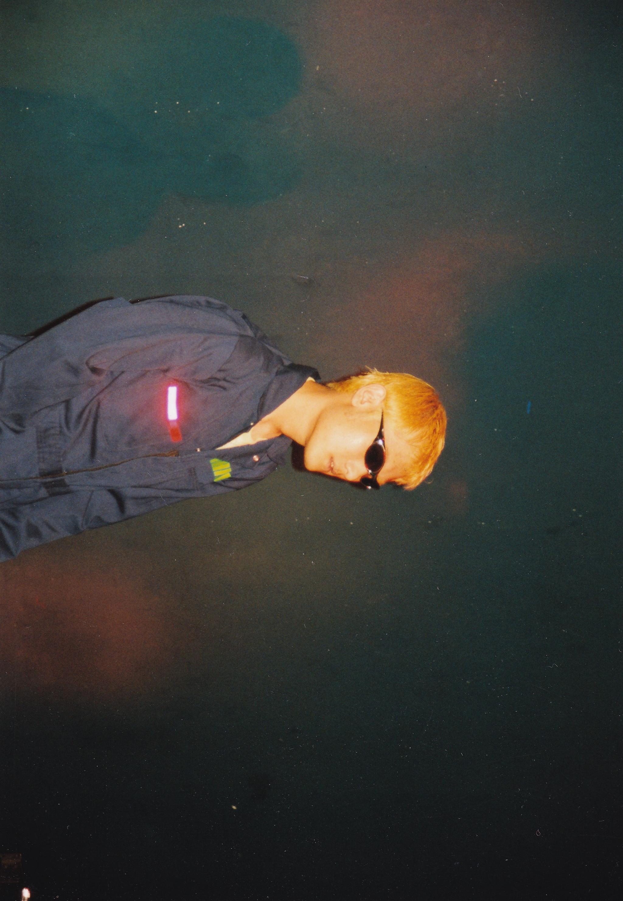Person with blonde hair in grey clothing lying on dark surface with red light on chest, green and brown tones in background.