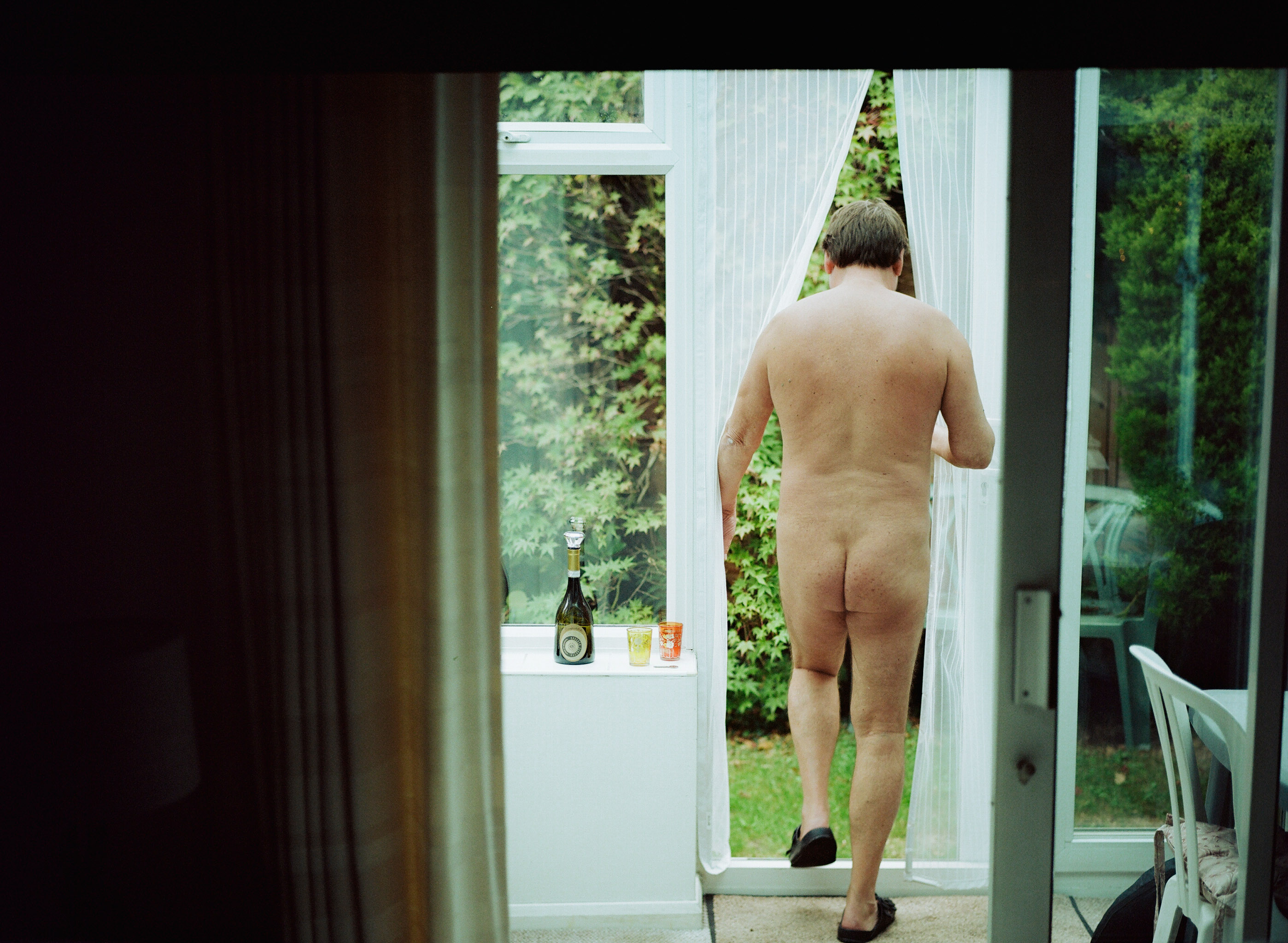 Naked man walking through open patio doors towards garden, wine bottle on windowsill, green trees visible outside.