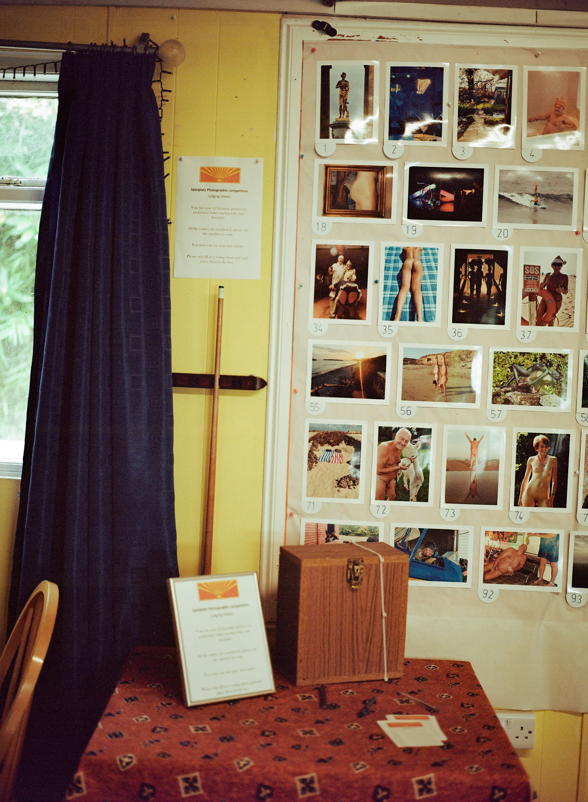 Interior with yellow walls, dark blue curtains, wooden stick, and white board displaying numbered colour photographs in grid layout.