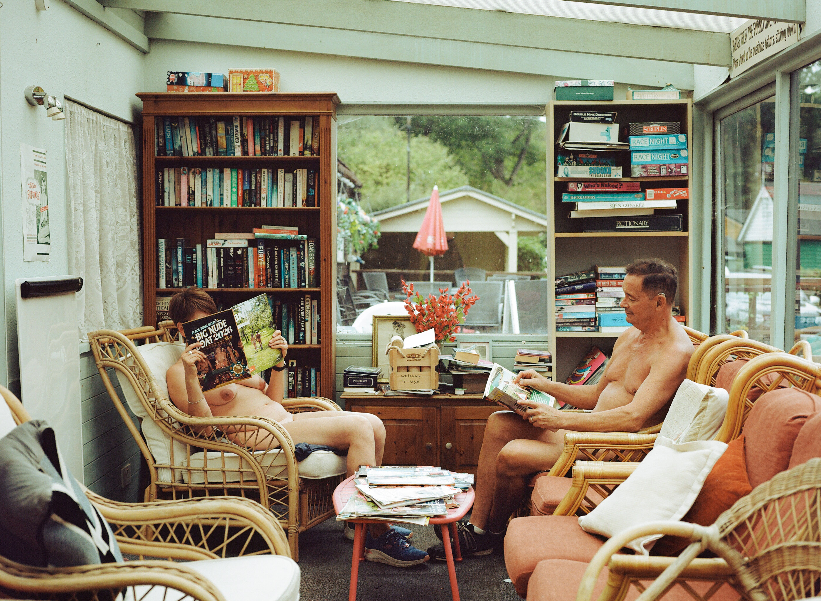 Two people reading in wicker chairs in glass-walled room with wooden bookshelves, papers scattered on small table, garden visible outside.