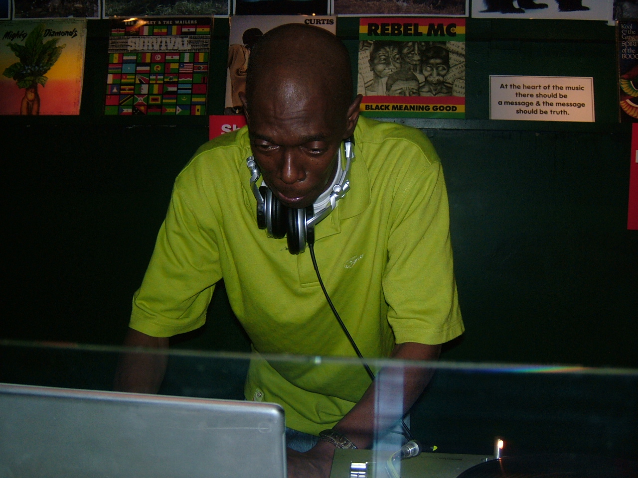 Black man in bright green shirt DJing with headphones at turntables, colourful posters on wall behind him.