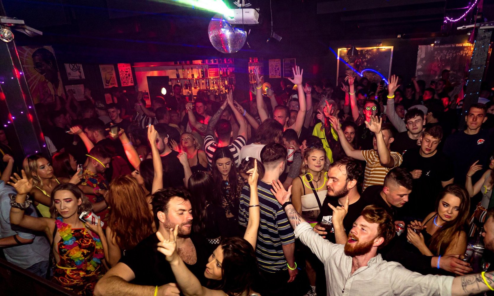 Crowded nightclub interior with people dancing, arms raised. Colourful lighting in purple, pink, and blue illuminates the scene.