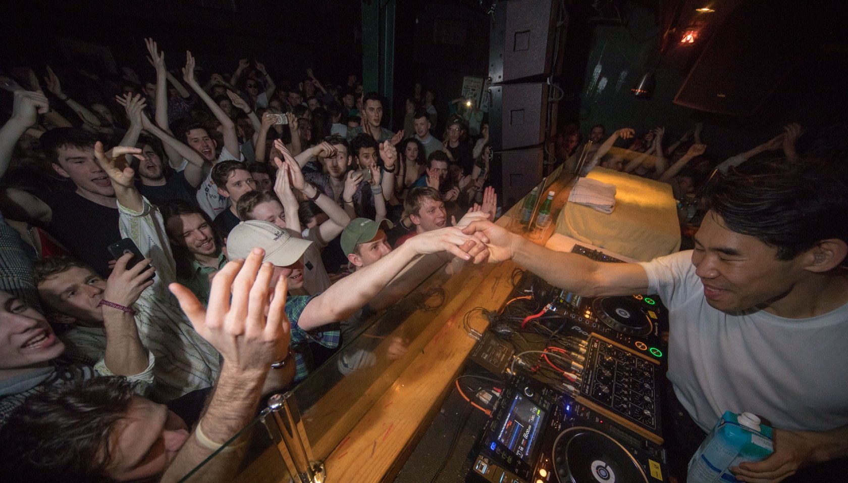 DJ performing at crowded nightclub with enthusiastic audience reaching towards the decks, orange lighting illuminating the scene.