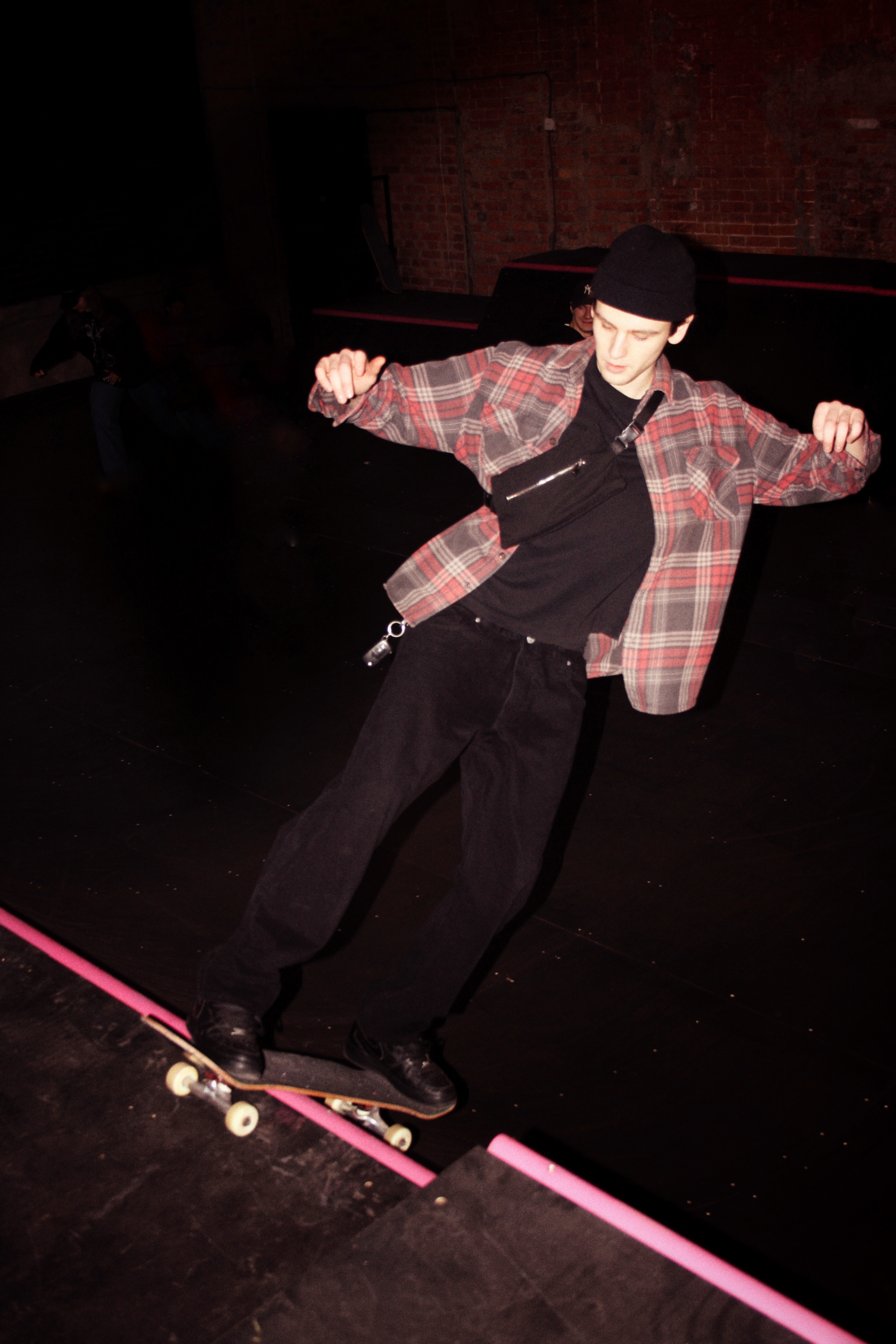A person wearing a plaid shirt and black trousers performing a trick on a skateboard in a dark setting.