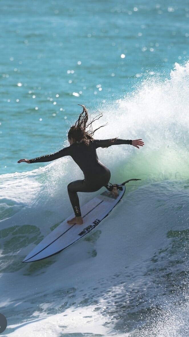 Surfer riding a wave on a blue surfboard, with arms outstretched, against a backdrop of turquoise water and sparkling light.