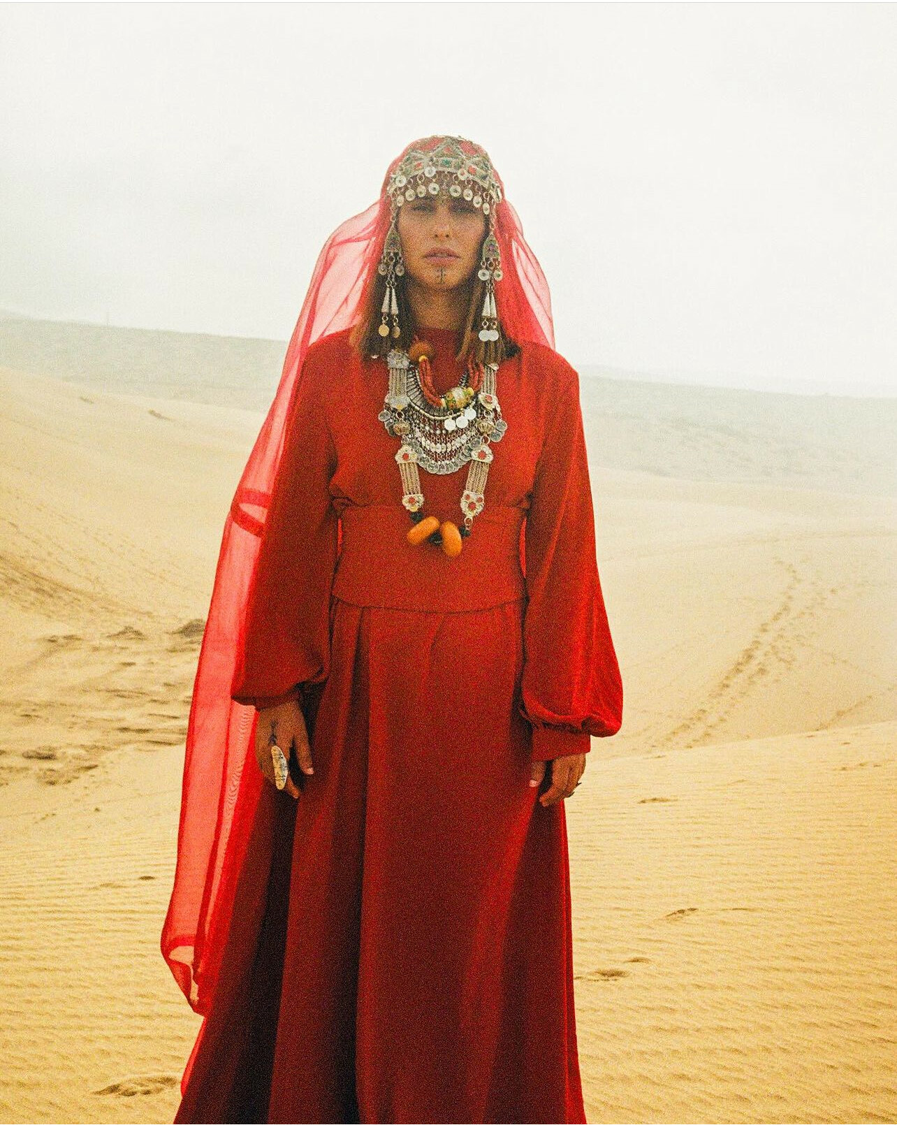 Ornate red dress with intricate patterned headpiece, gold jewellery, and flowing scarlet veil in desert landscape.