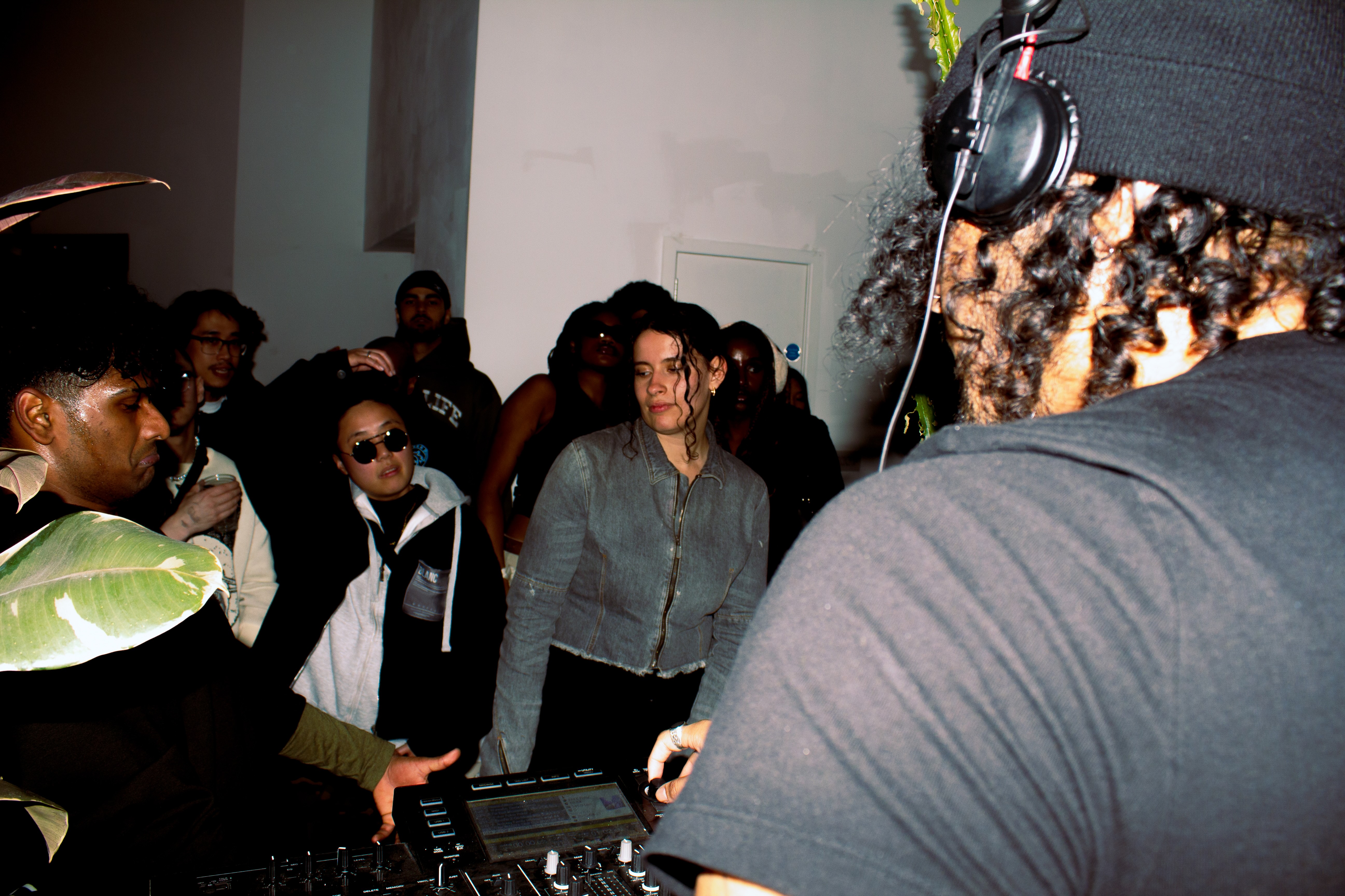 Group of people gathered around DJ equipment in dimly lit room, with DJ in foreground operating mixing deck whilst crowd watches.