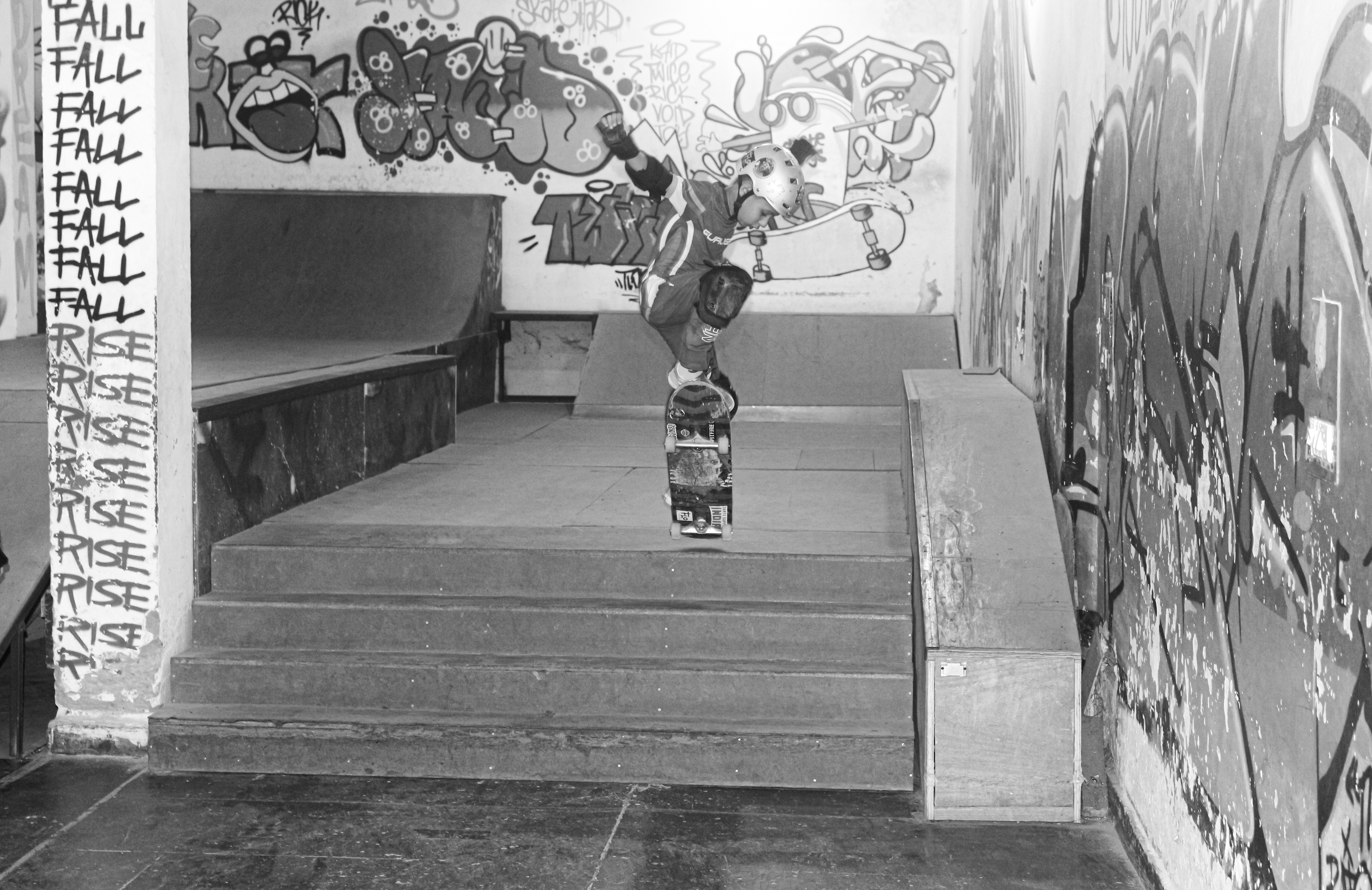 Black and white image showing skateboarder on concrete steps with graffiti-covered walls. Repeated "FALL" text visible on left wall.
