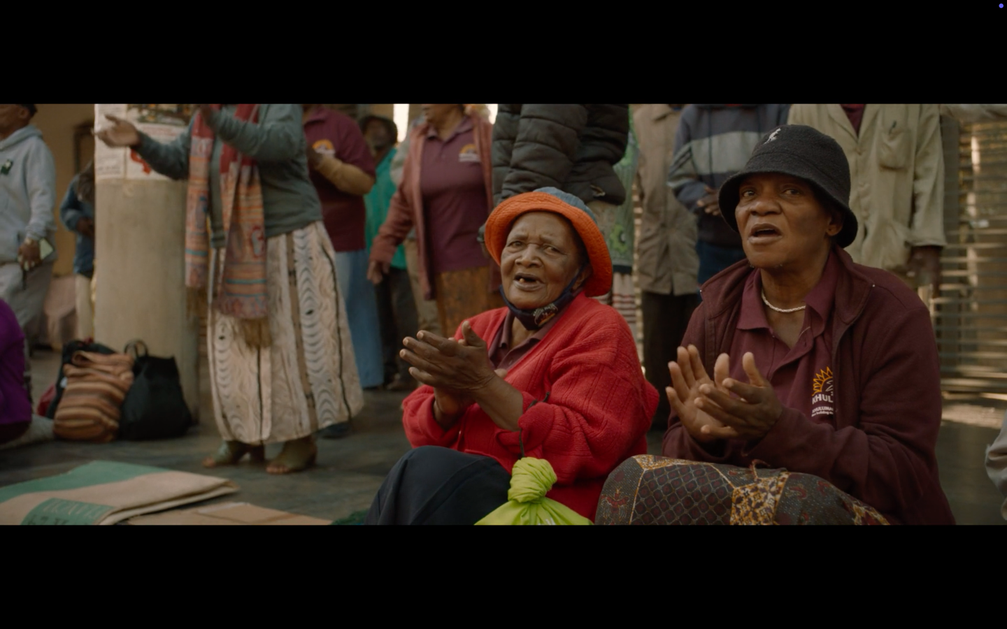 Two elderly women wearing traditional clothing and hats, sitting on a bench and clapping.