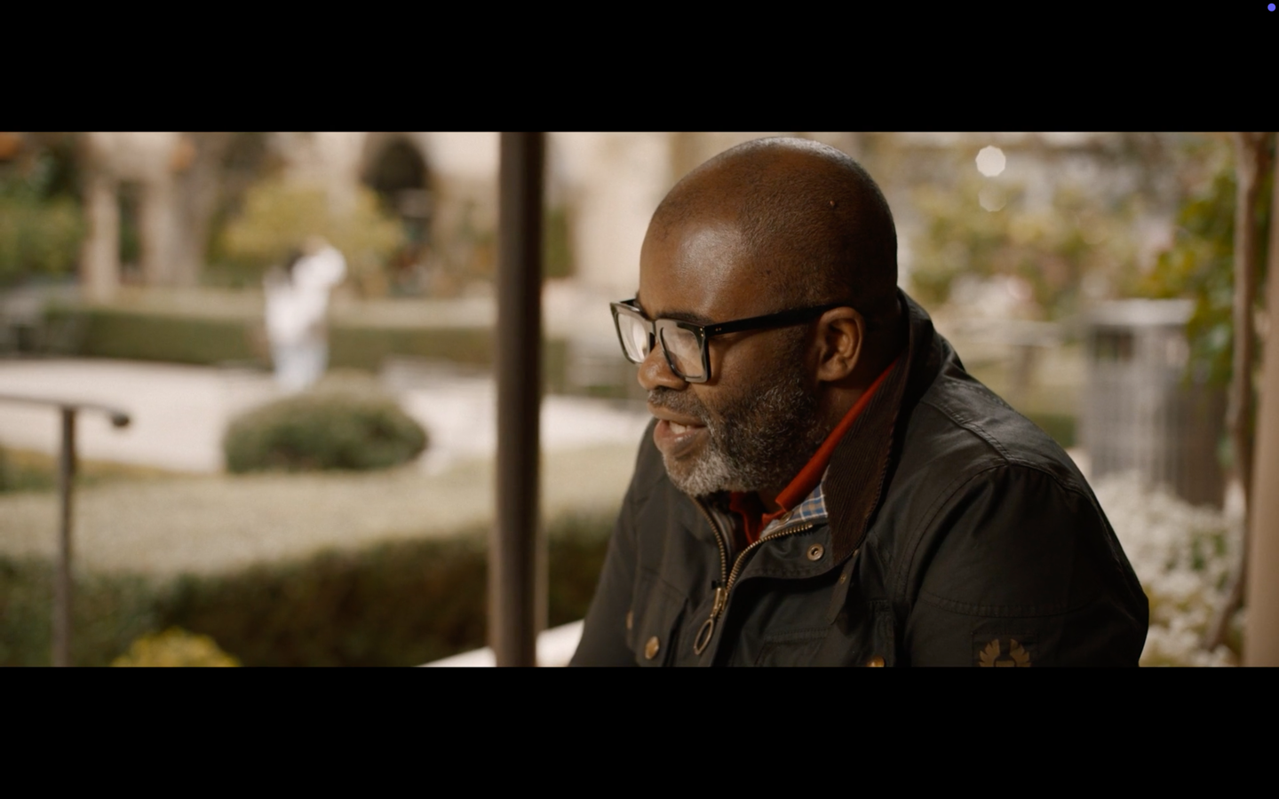 A bearded man in glasses wearing a dark jacket and red scarf stands in a garden setting.