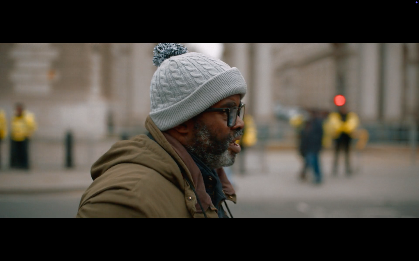Man in a grey woollen hat and coat, walking on a city street in the cold.