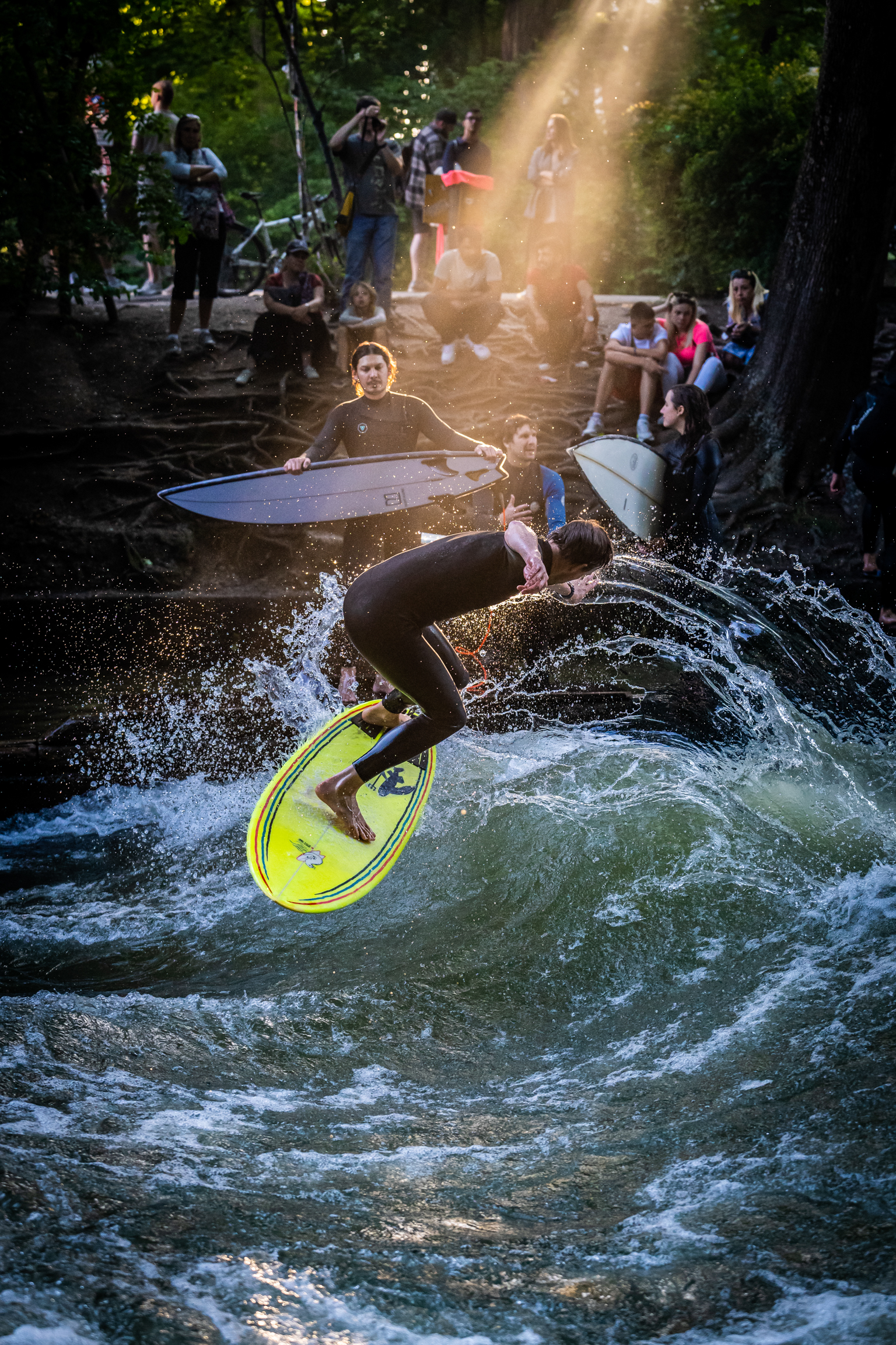 Person in wetsuit riding yellow paddleboard on rushing river water, with spectators watching from wooded banks above.