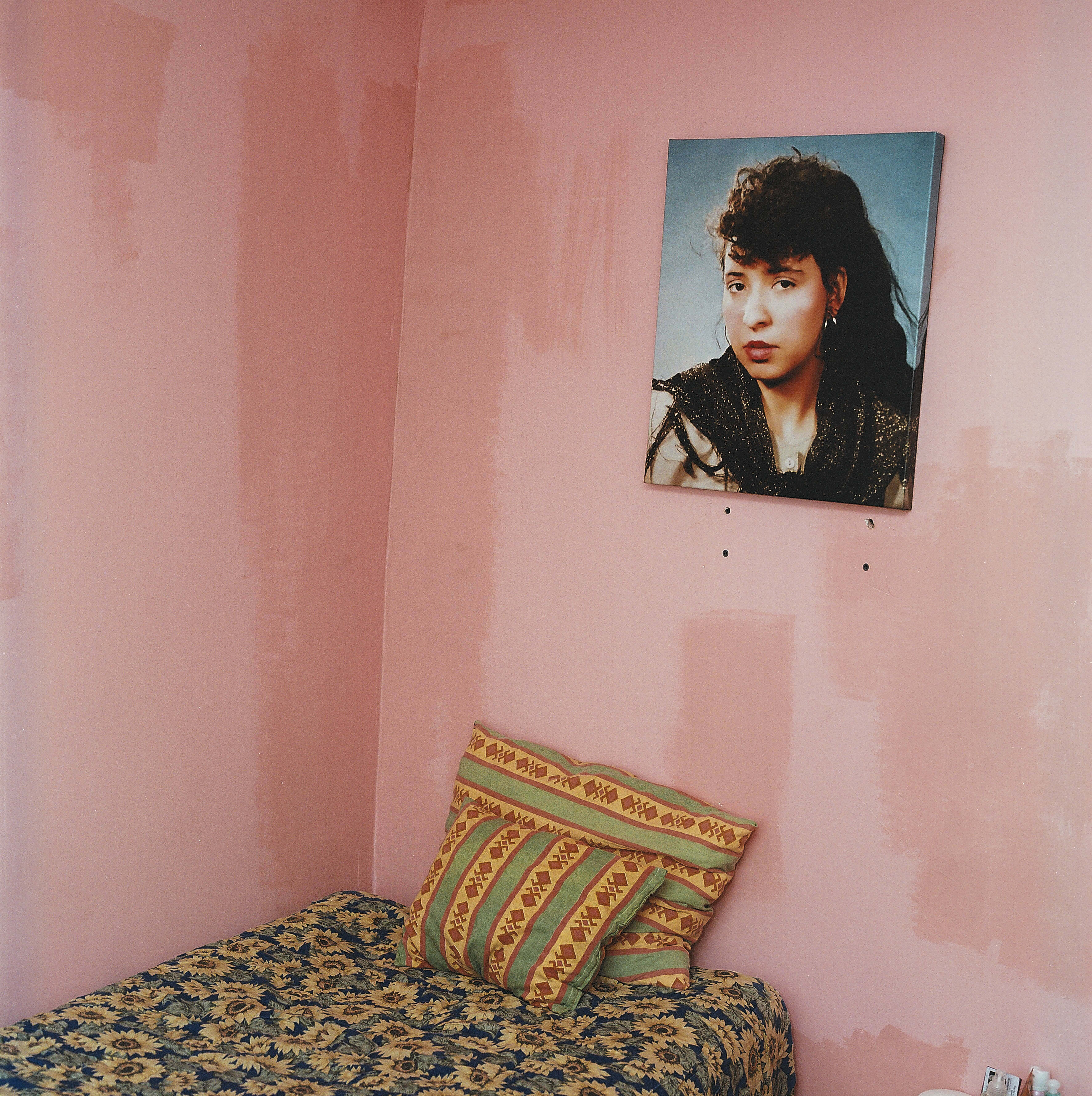 A portrait of a person hangs on a pink wall, with a patterned cushion on the bed in the foreground.