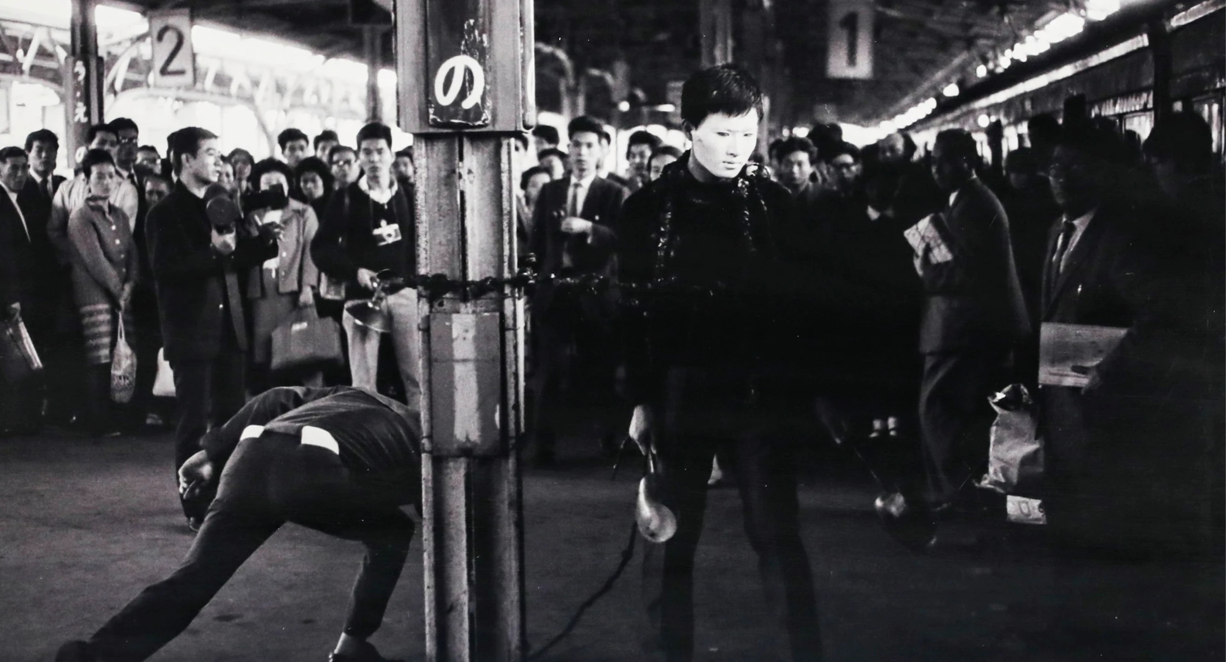Crowded urban street at night, people gathered on platforms of train station, silhouettes and shadows cast, focus on central figure in motion.