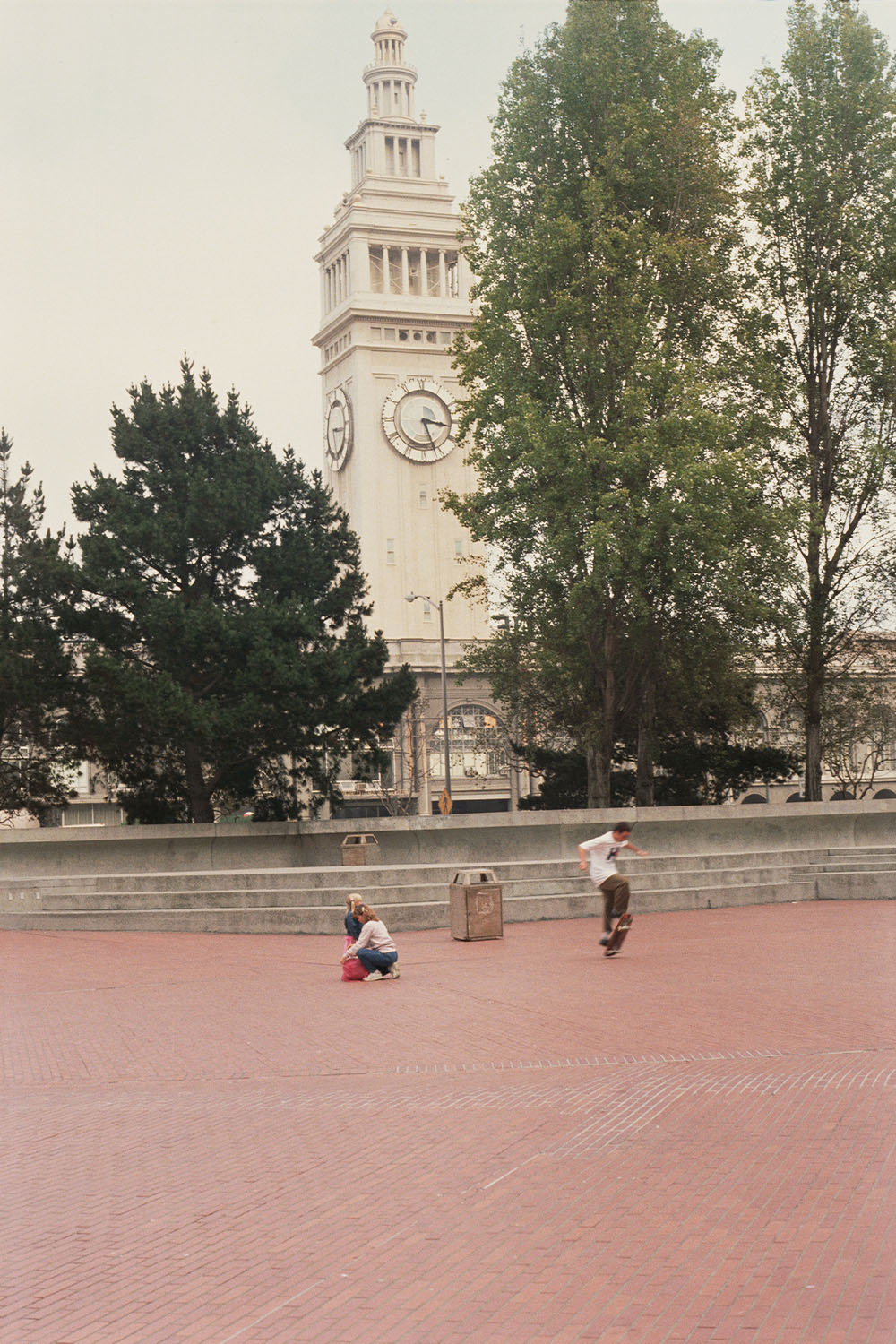 White clock tower with columned sections rises behind green trees. Two people sit on brick plaza with concrete steps and bollards.