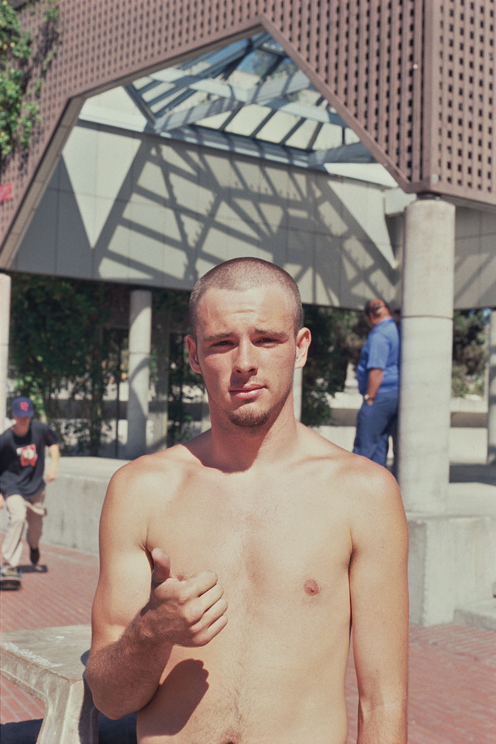 Shirtless man with short hair giving thumbs up outdoors, modern glass and steel canopy structure with geometric patterns behind him.