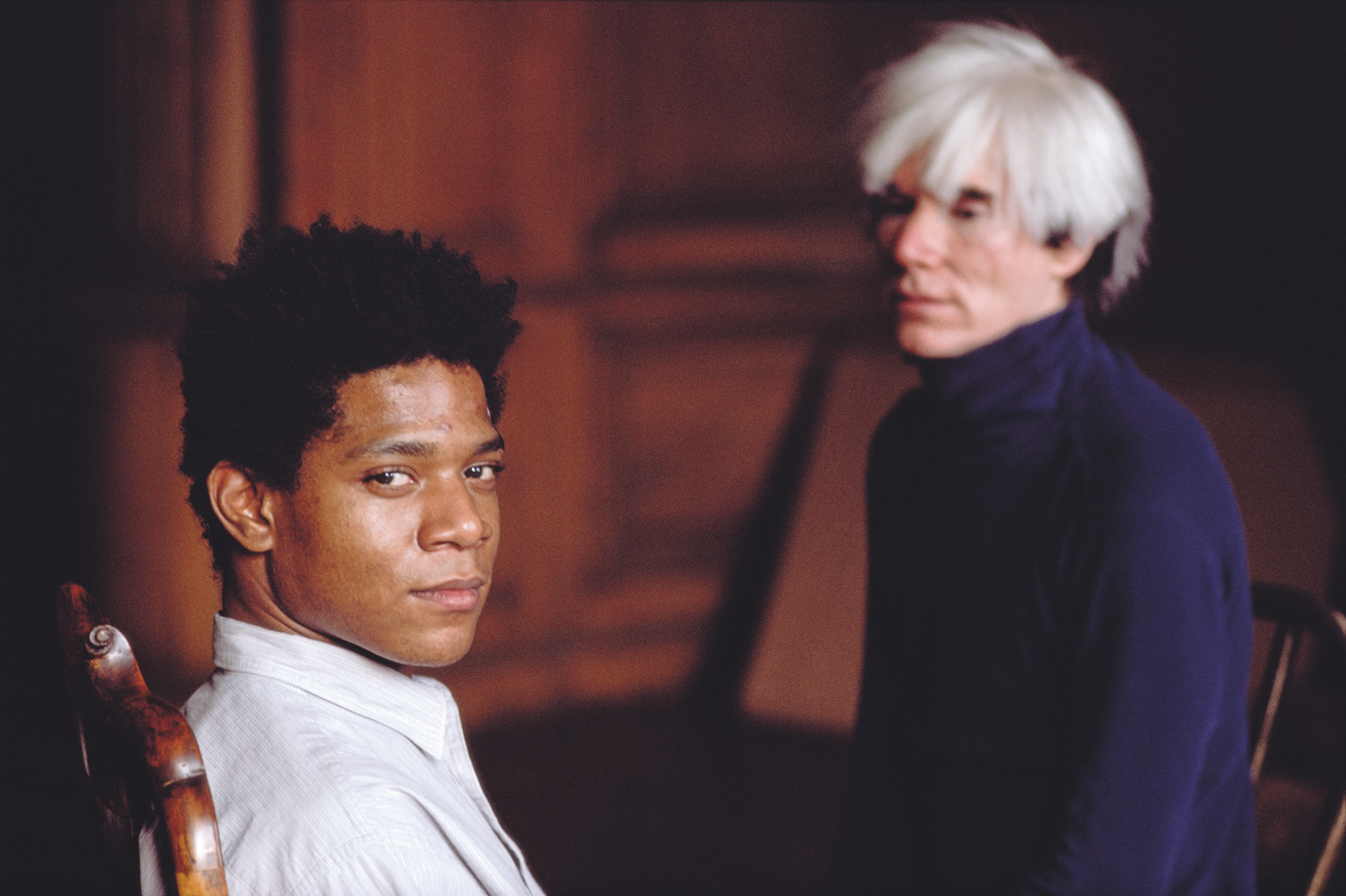 Young Black man in white shirt sits beside older white-haired man in dark jacket against warm reddish-brown background.