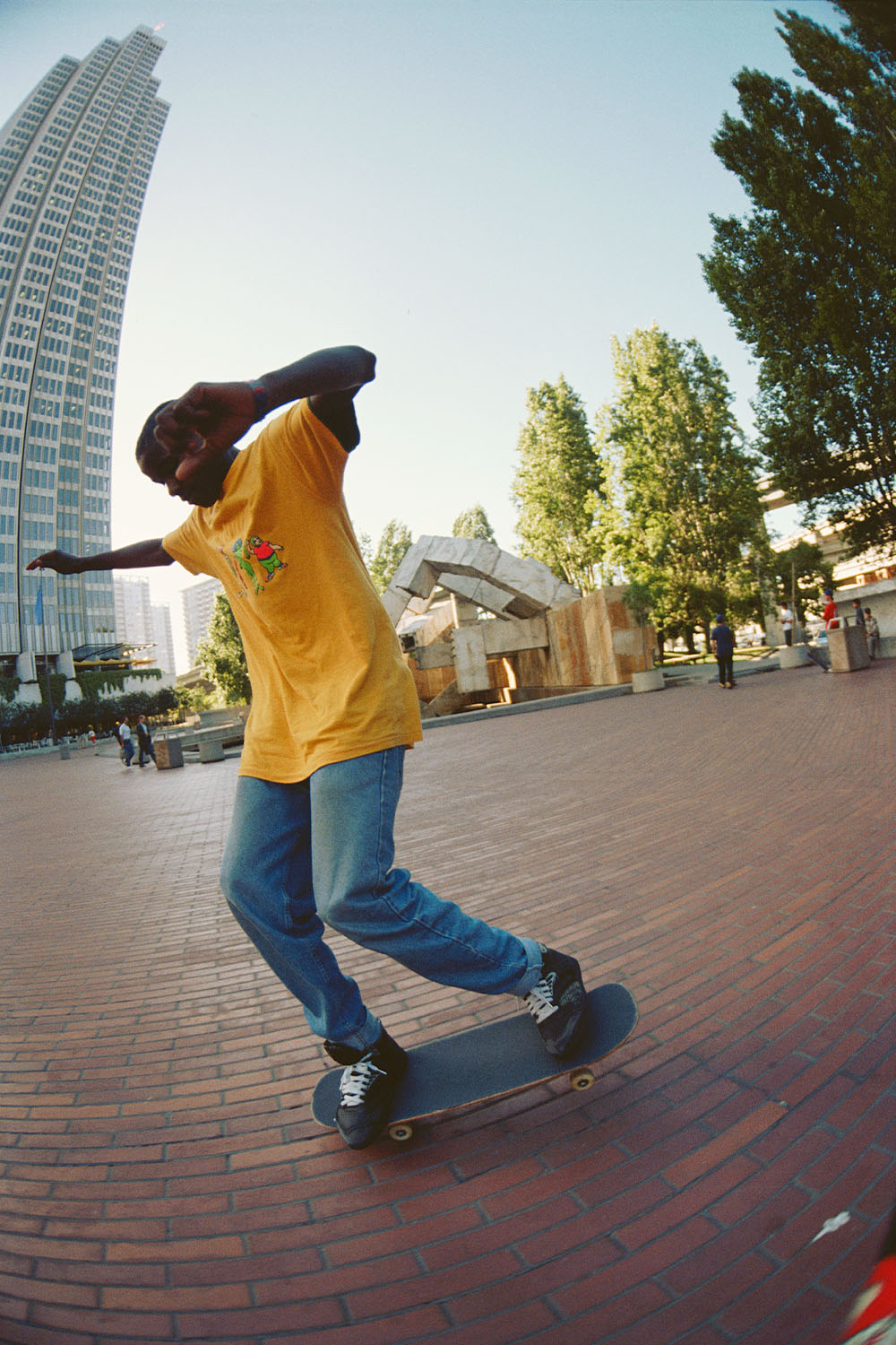 Person skateboarding on brick plaza wearing yellow t-shirt and jeans, with tall white building and trees in background.