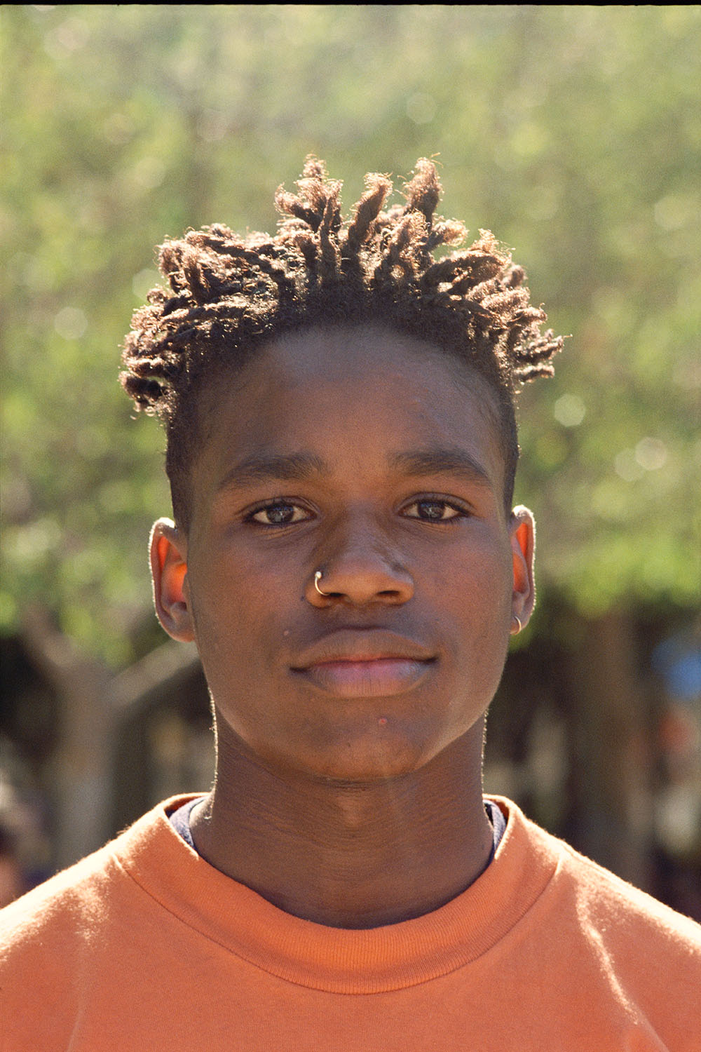 Young Black man with blonde locs wearing orange shirt, nose piercing visible, green foliage blurred in background.