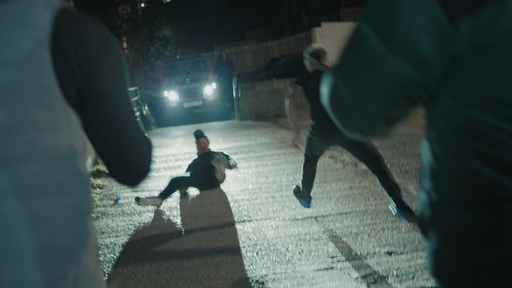 Person kneeling on wet pavement at night, viewed through car windscreen with bright headlights illuminating the scene.