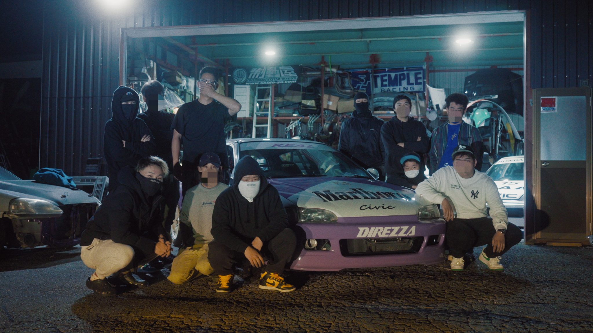 Group of people posing with modified cars outside illuminated garage at night, wearing dark clothing and face coverings.