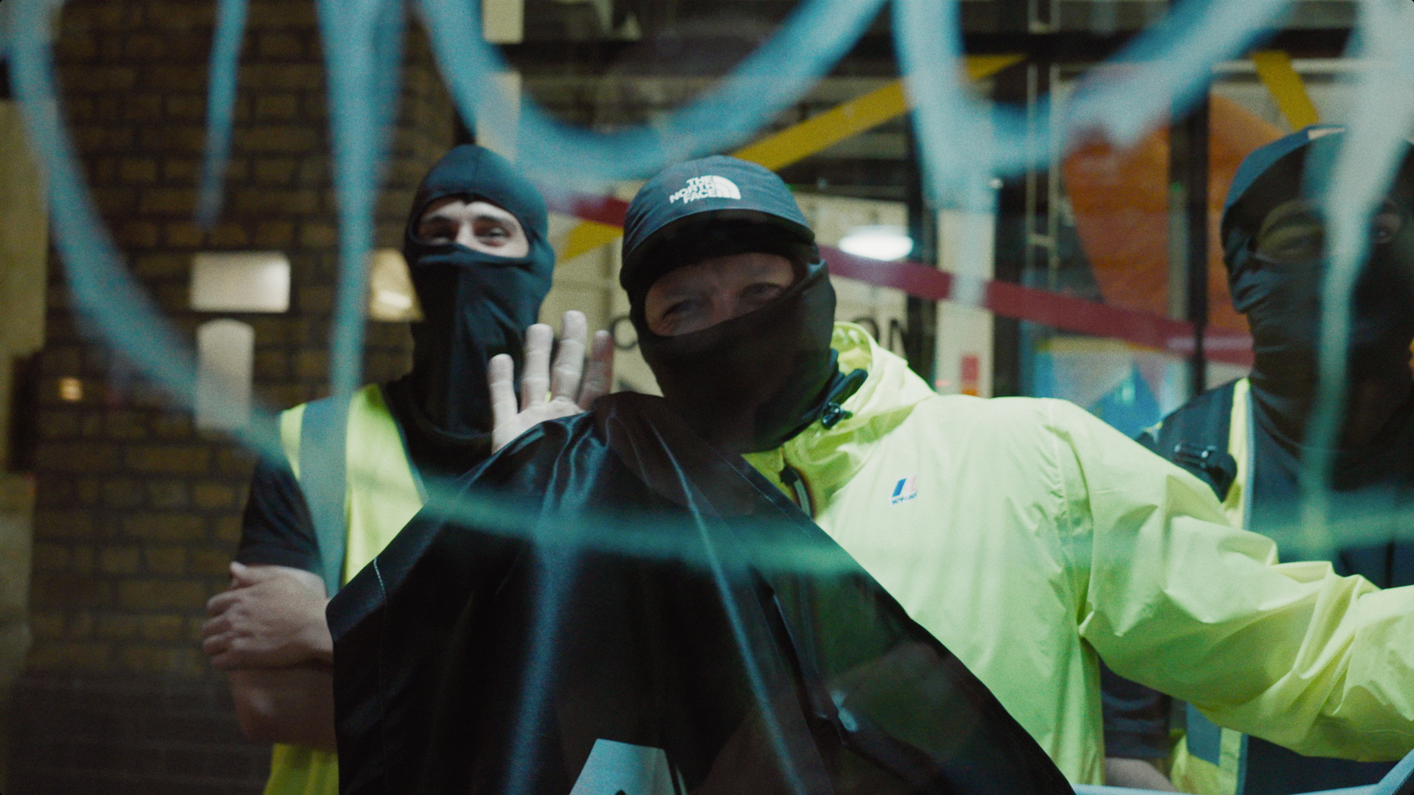 Two people in black face coverings and caps, one wearing bright yellow jacket, standing behind turquoise metal bars or railings.