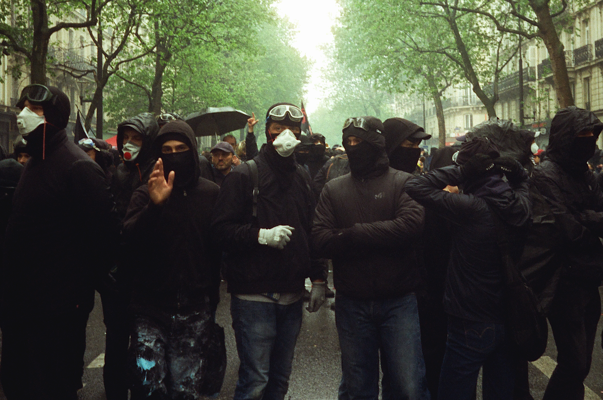 Crowd of people in dark clothing and face coverings gathered on tree-lined street with green foliage and buildings visible.