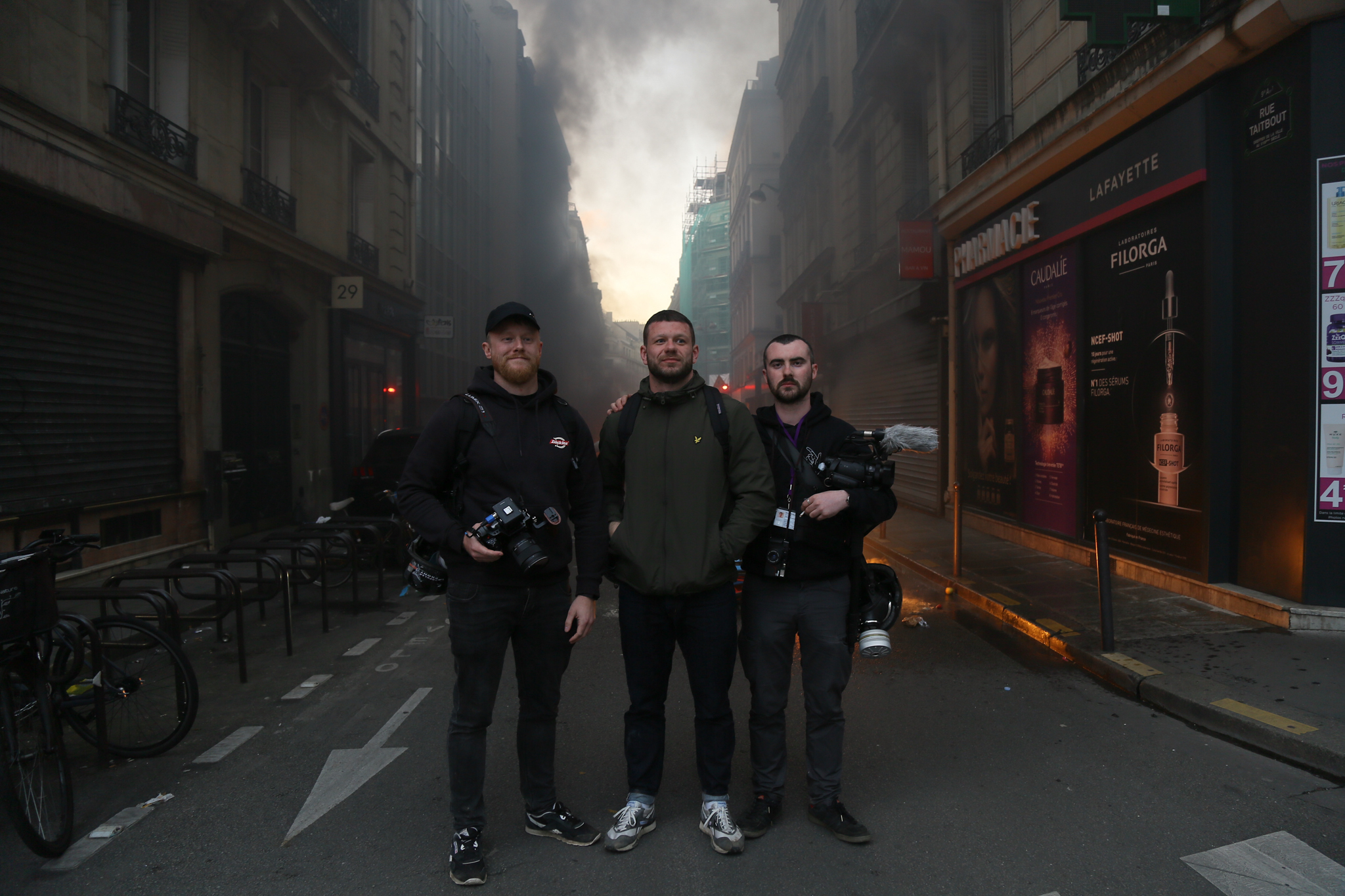 Three men in dark clothing standing on narrow street with tall buildings, motorcycles parked nearby, overcast sky visible above.